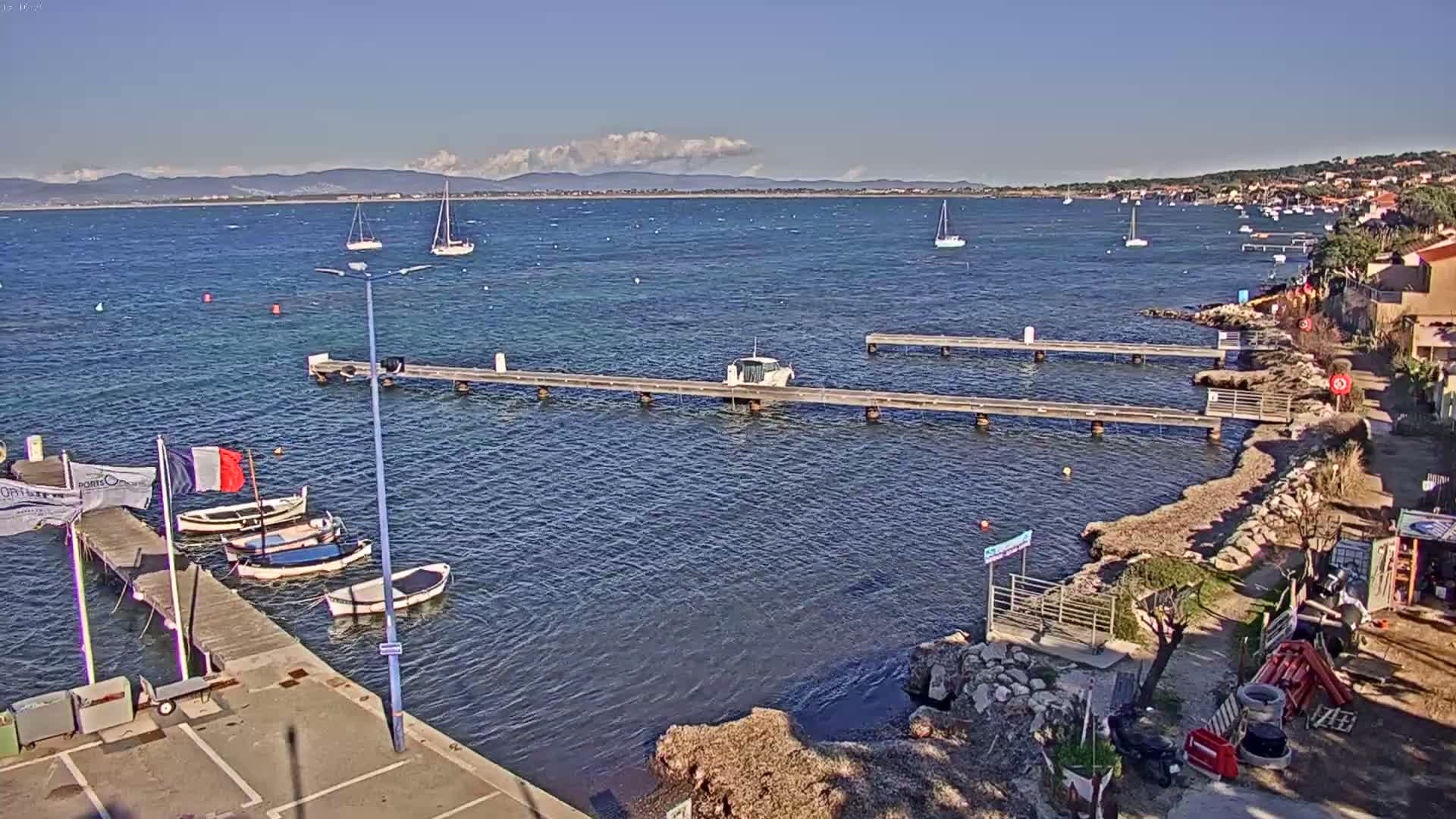 A calm, sunny day reveals a waterfront scene with several small boats moored at wooden piers, a larger boat in the water beyond the piers, and a distant shoreline with buildings under a partly cloudy sky.