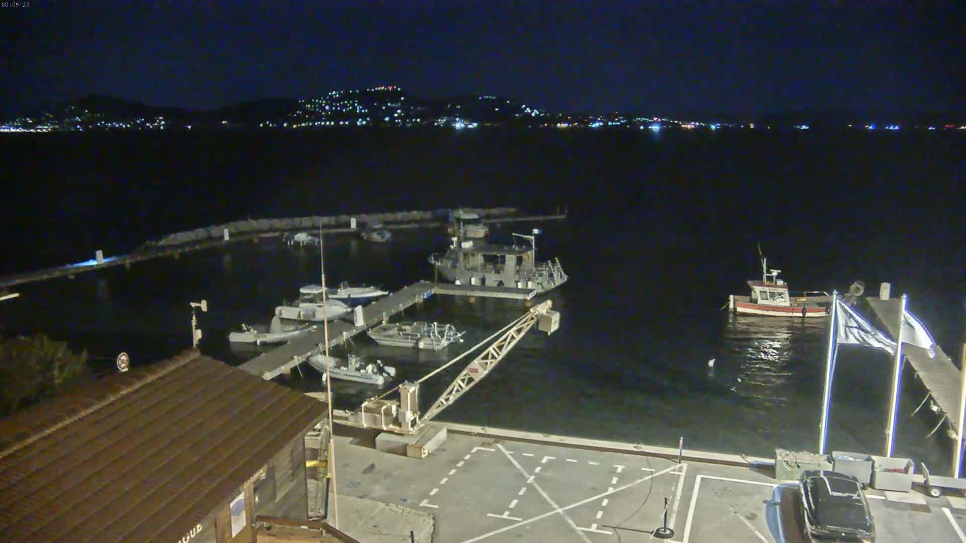 A nighttime view of a harbor with several boats docked, a small crane, and a distant city skyline visible across the water under a clear, dark sky.
