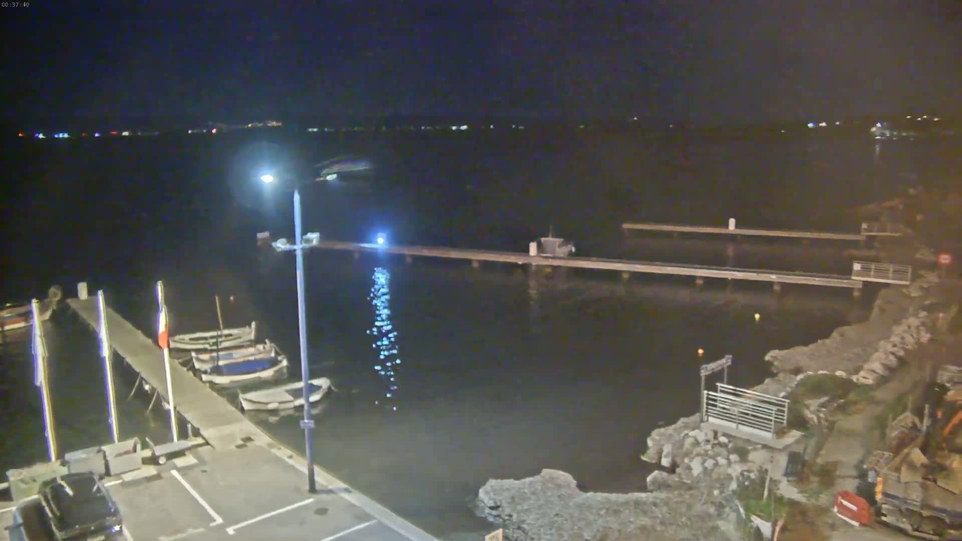 At night, several small boats are moored at a dock extending over dark water, with lights illuminating the scene.