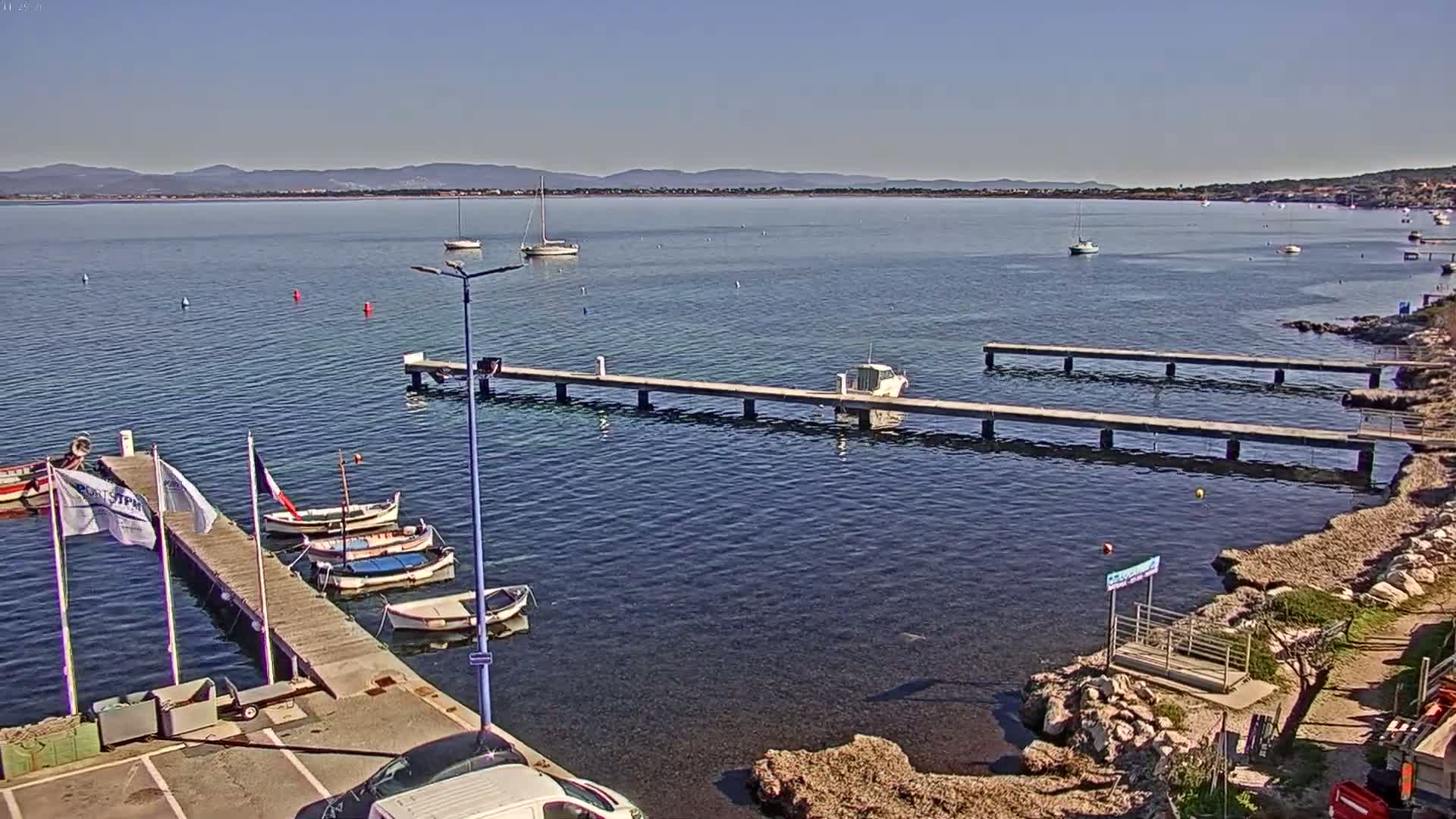 A calm, sunny day reveals a harbor scene with several boats moored at long piers extending from a rocky shoreline, with a distant mountain range visible in the background.