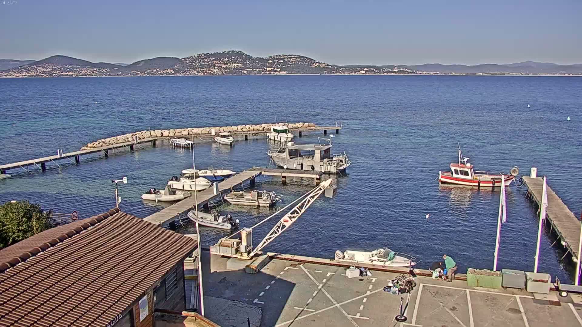 A small harbor filled with various boats and a crane, under a clear sunny sky, with a mountainous coastline visible in the distance.