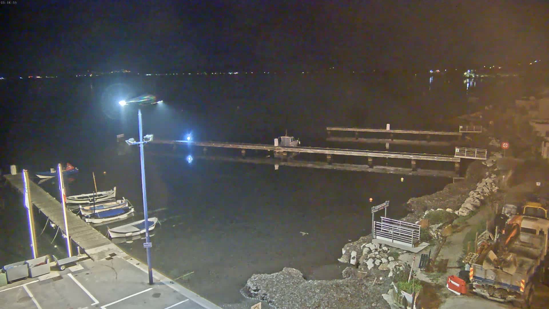 At night, several small boats are moored at a dock next to a body of dark water, with a small construction vehicle visible on the shore.