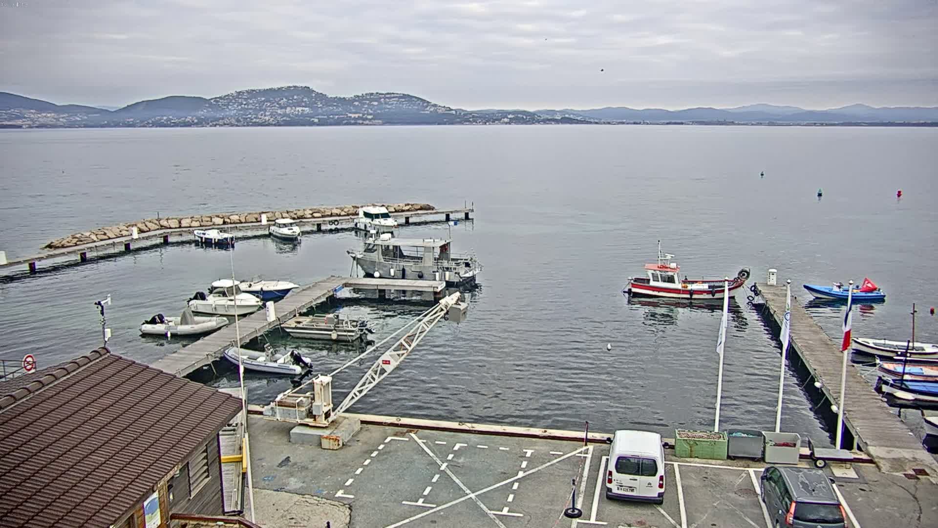 A small marina with several boats docked at piers, a calm body of water extending to distant hills under an overcast sky.