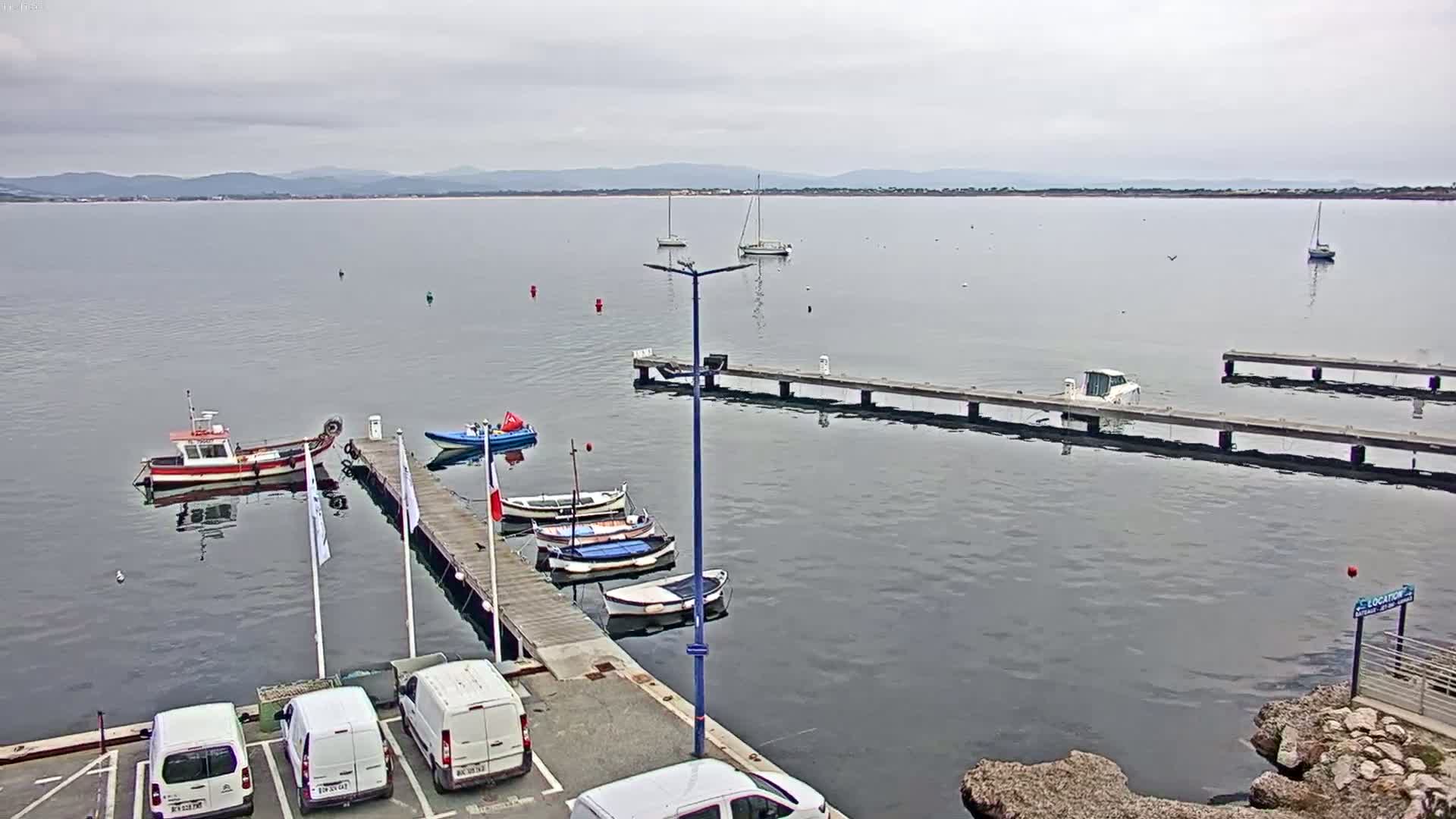 Several boats are moored at a dock on calm, gray water under an overcast sky, with additional boats and sailboats visible further out in the bay and mountains in the far distance.