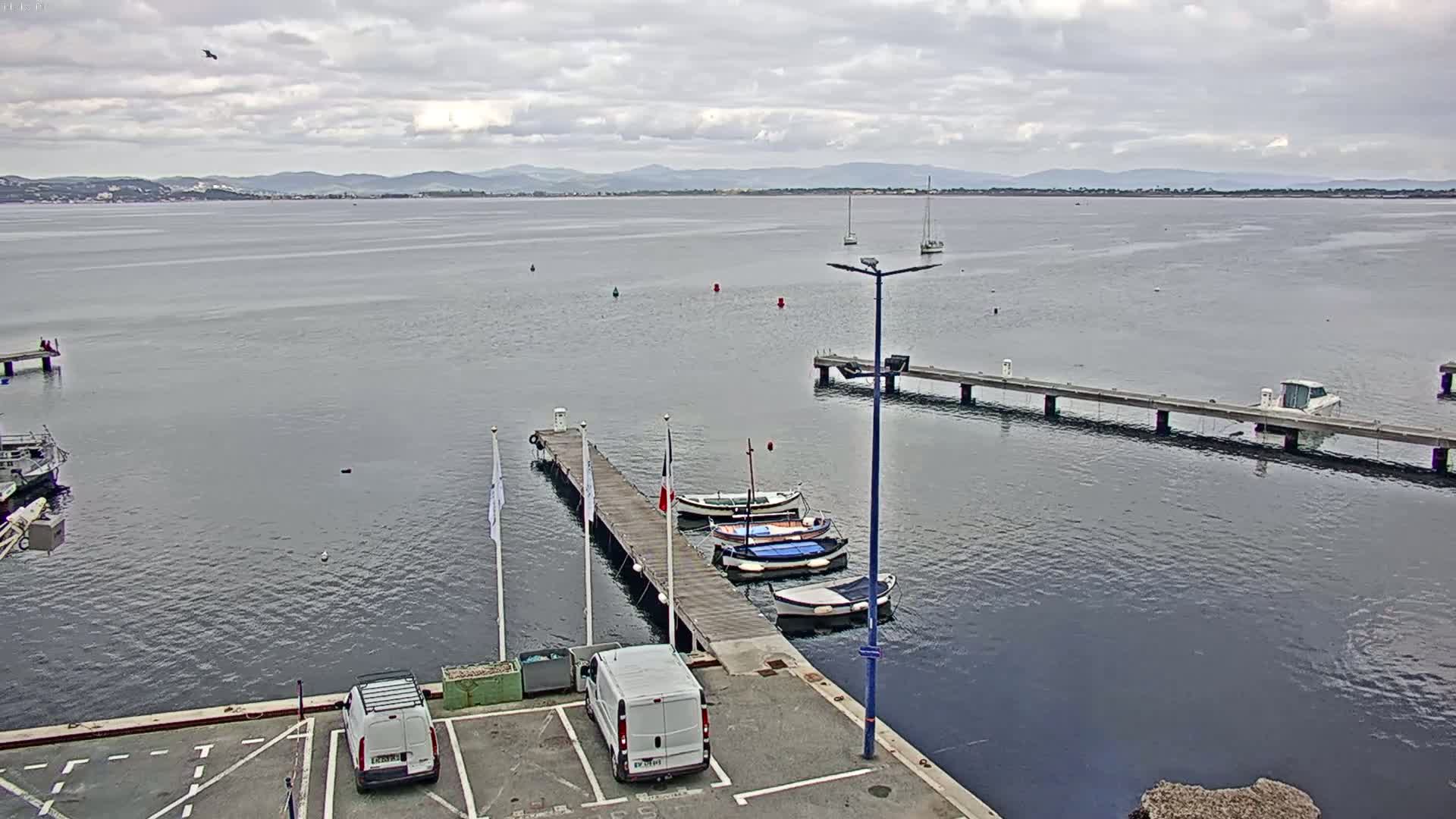 A calm body of water under a cloudy sky is seen from a high vantage point, showing several small boats moored at a pier with two vans parked nearby.
