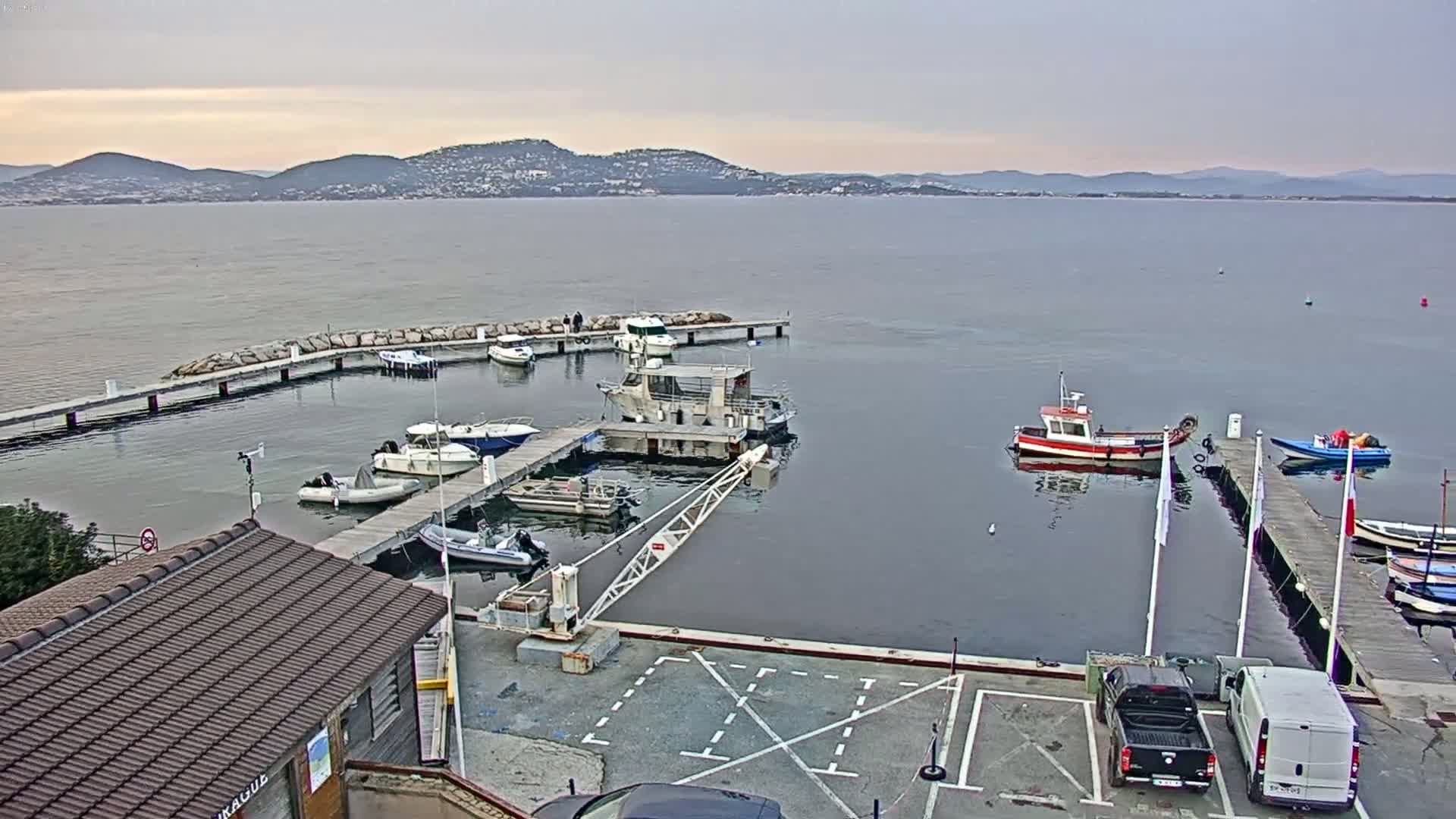 A small marina with several boats docked at piers, a small building with a brown roof, and a few vehicles in a parking lot, all under a cloudy sky with distant hills visible across calm water.