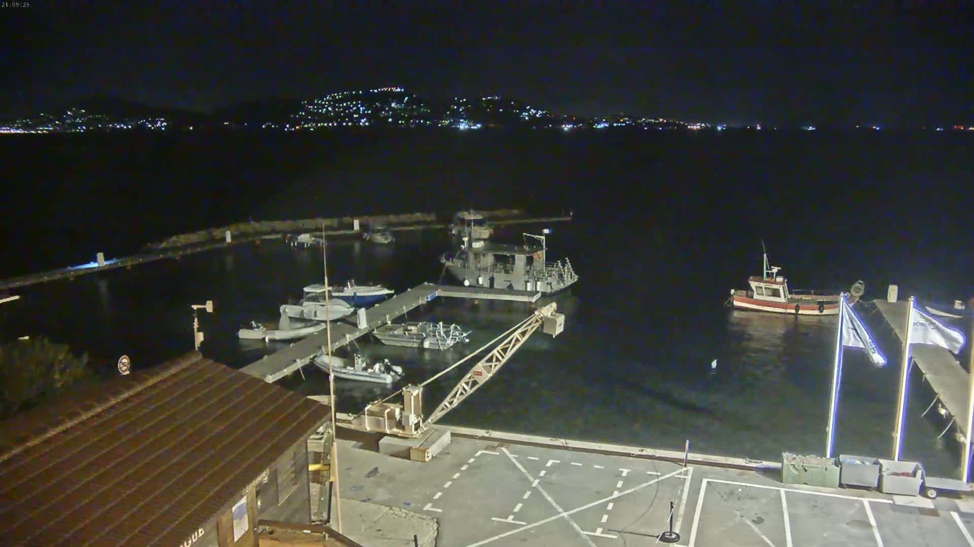 A nighttime view of a marina with several boats docked, a small crane, and a distant city illuminated on a hillside across the water.