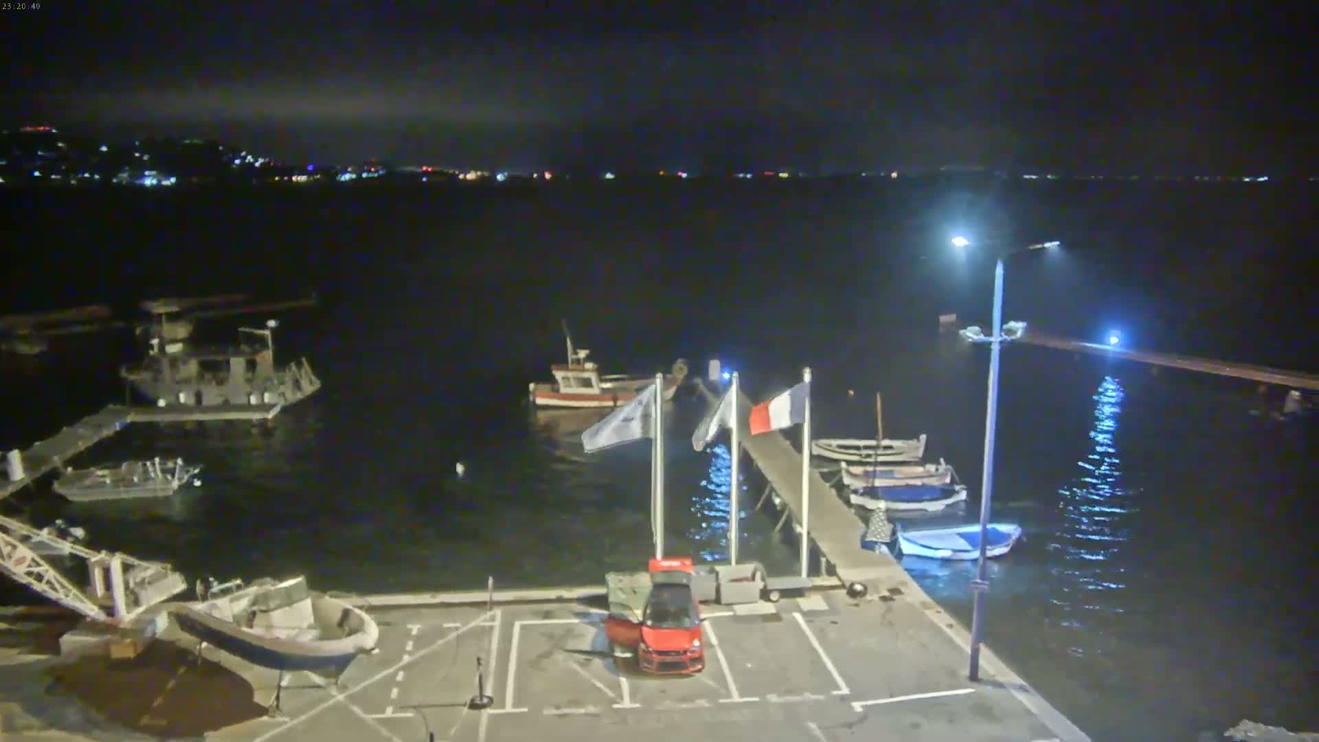 A small harbor at night with several boats moored at a pier, a red car parked nearby, and distant city lights visible across the calm water.