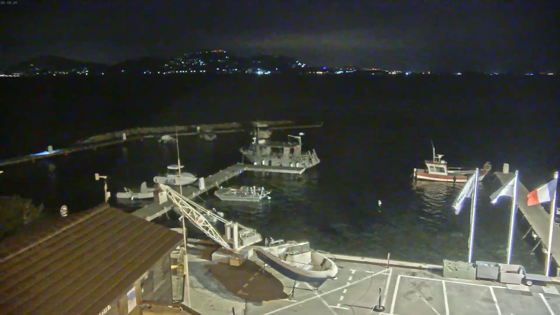 A nighttime view of a harbor with several boats moored at docks, under a dark, mostly clear sky, with a distant town illuminated on the horizon.