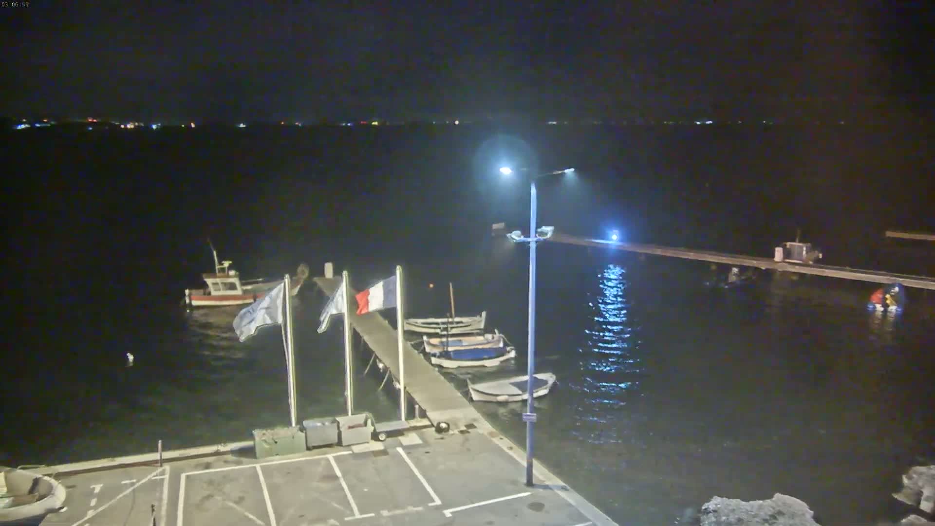 At night, several small boats are moored at a dock illuminated by a lamppost, with calm water reflecting light and a distant shoreline visible.