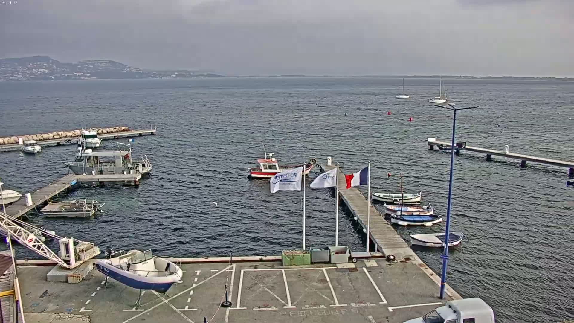A small harbor with several boats docked at piers, under an overcast sky, overlooks calm water and a distant shoreline.