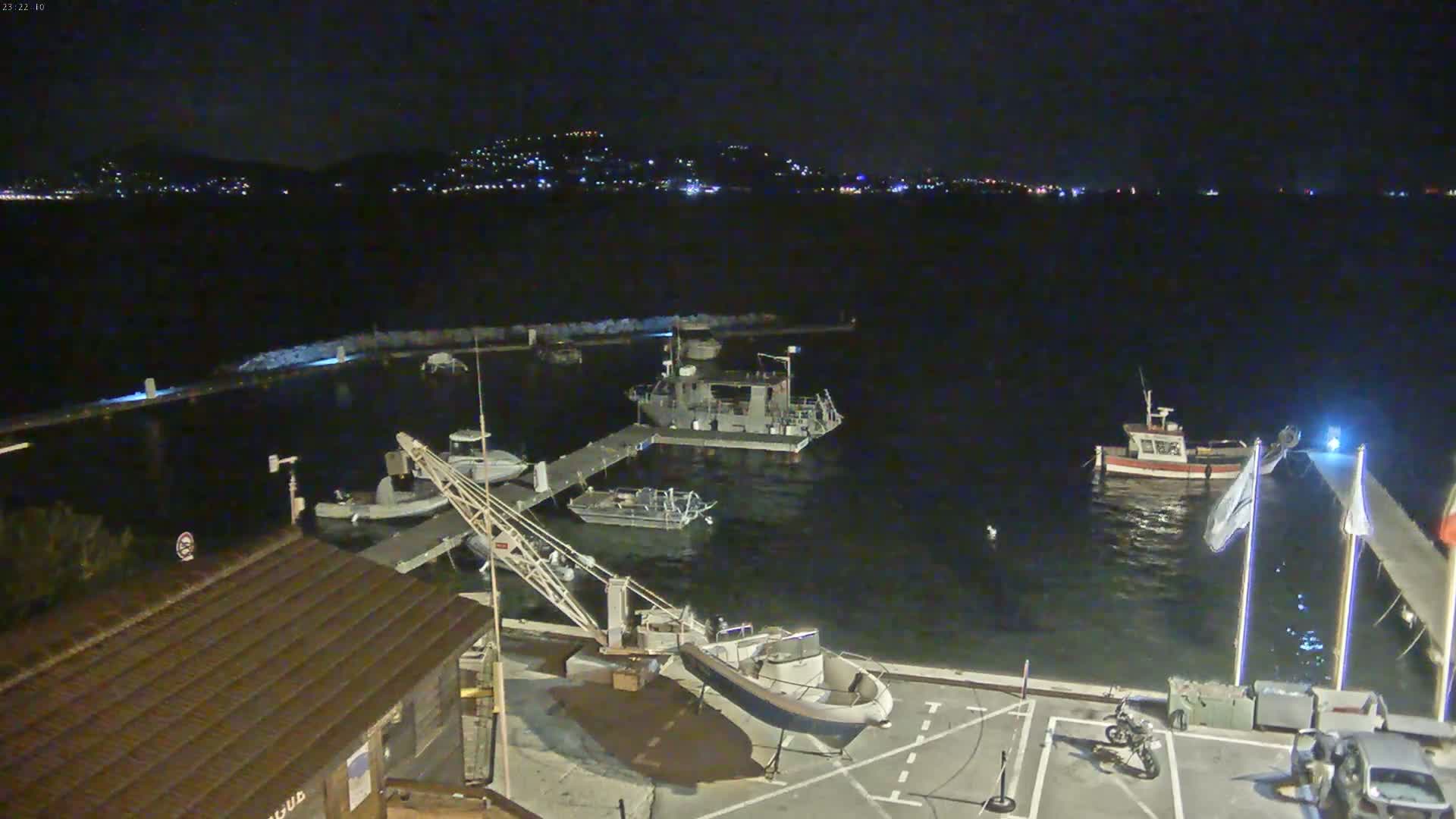 A nighttime view of a harbor with several boats docked, a crane, and a building, under a clear, dark sky with a city skyline visible in the distance.