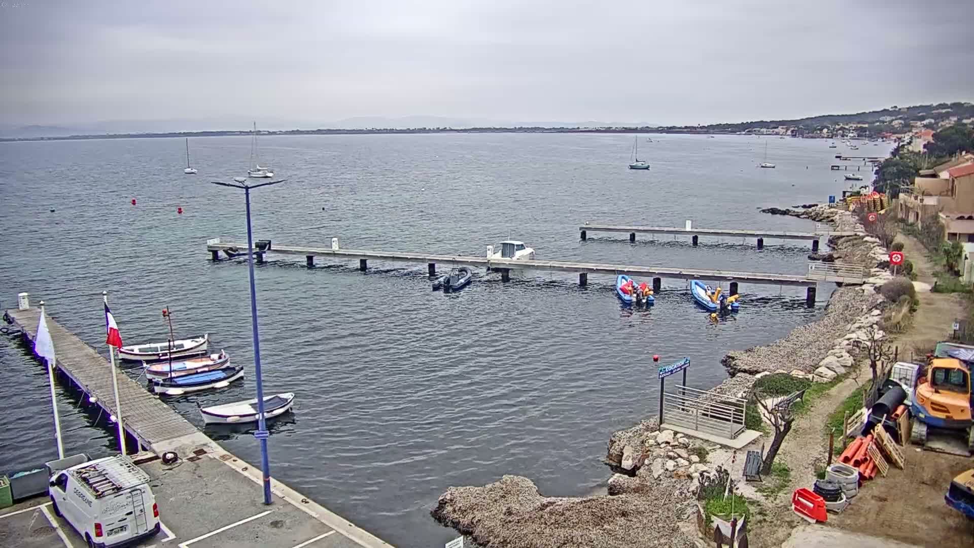A calm body of water under an overcast sky features several small boats moored at multiple piers along a rocky shoreline.