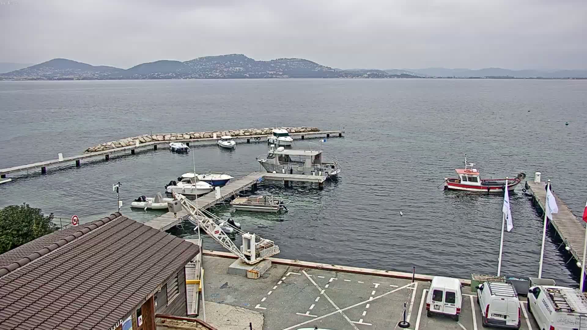 A small marina with several boats docked at piers, a crane, and a building, overlooks calm water under an overcast sky, with distant mountains visible on the horizon.