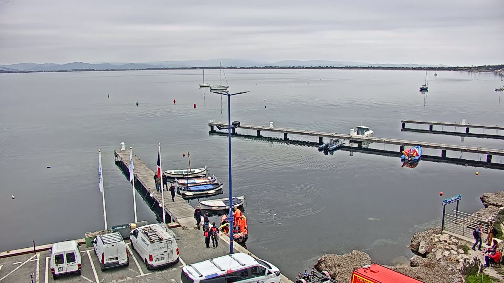A calm, overcast day shows a harbor with several boats docked at wooden piers, where people are gathered near a small rescue boat.