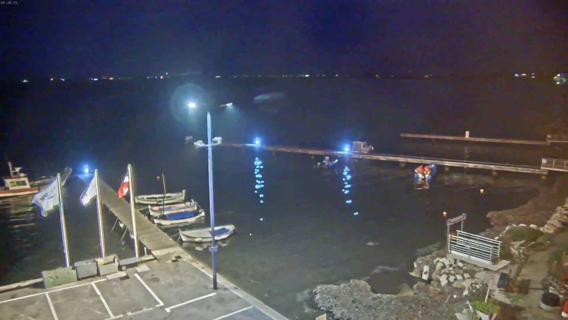 At night, several boats are moored at a pier next to dark water, and people are visible near the water's edge.