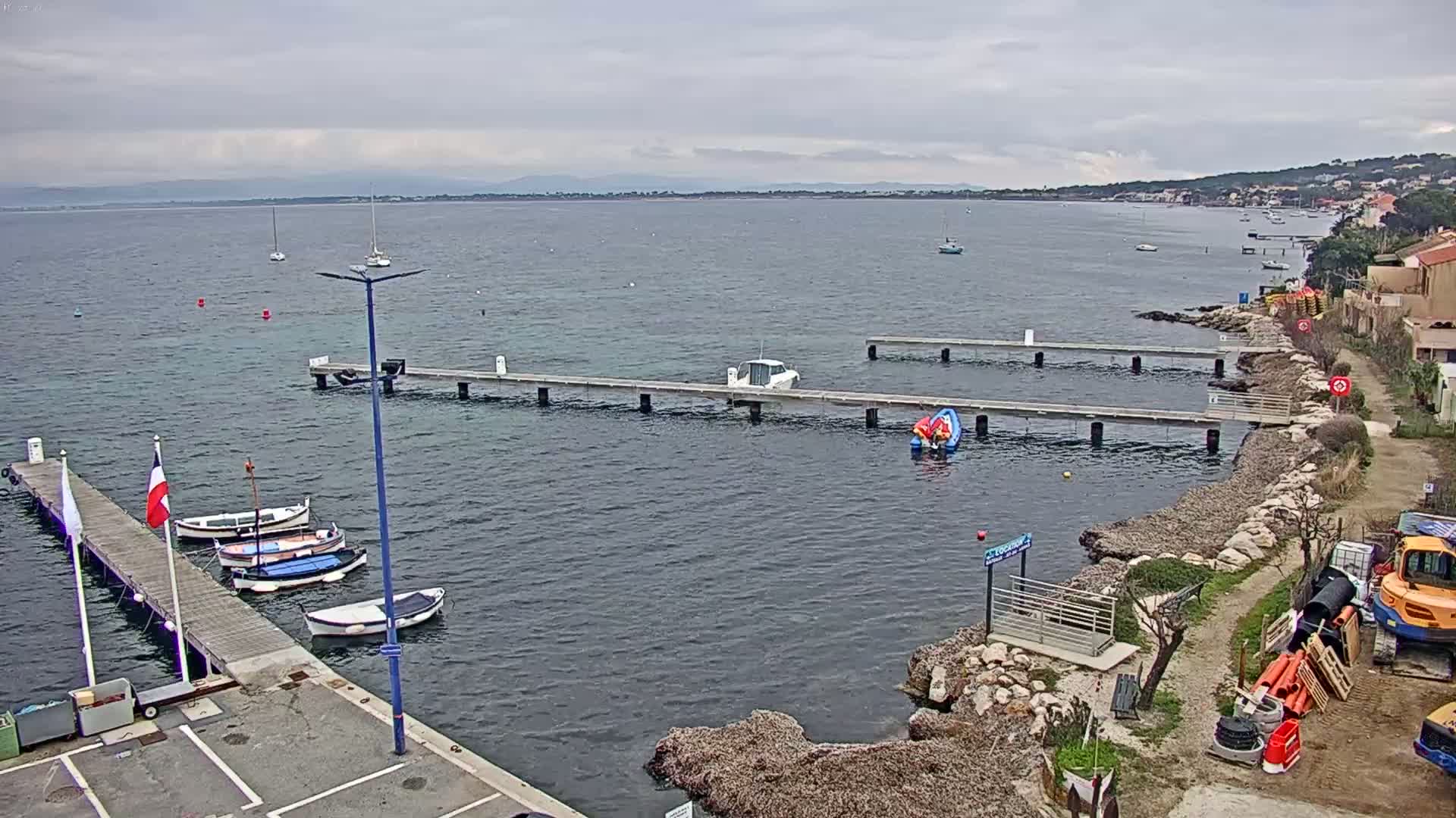 A calm, overcast day reveals a small harbor with several boats docked at piers, extending into a body of water with a distant shoreline visible under a cloudy sky.