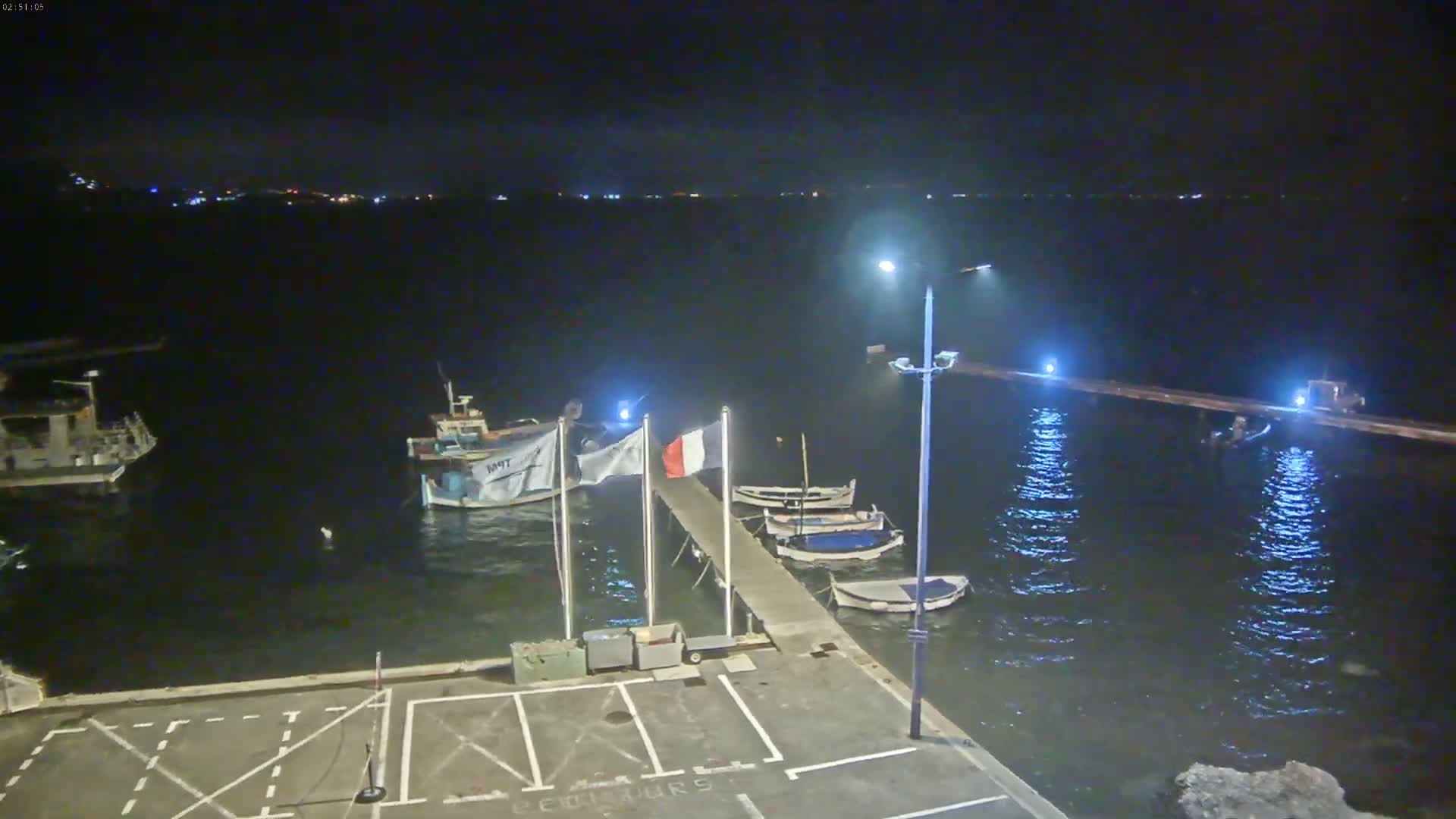 At night, several small boats are moored at a pier next to a parking lot, under a calm, dark sky.