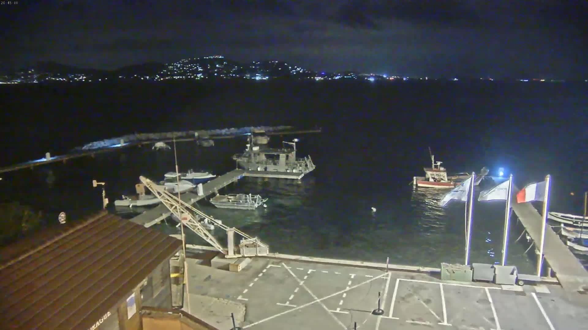 At night, several boats are moored at a marina, with a city’s lights visible across the water.