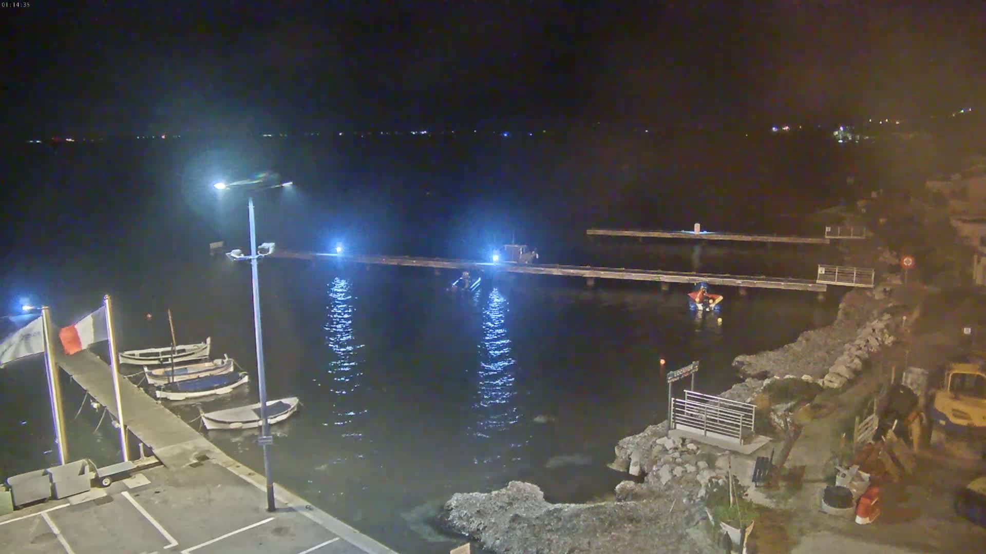 A nighttime view of a calm body of water with several small boats docked at a pier, illuminated by lights along the pier and a streetlight on land.
