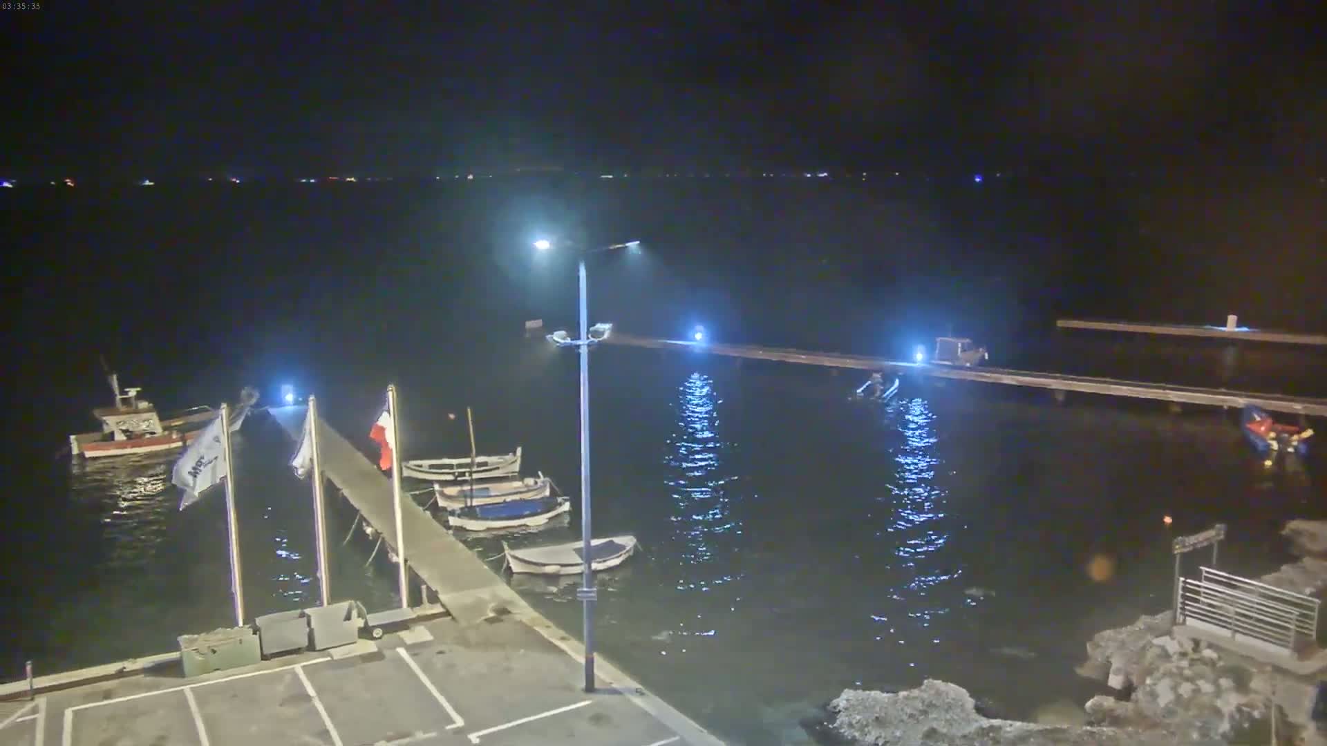 Several small boats are moored at a pier at night, with a vehicle parked on the pier and calm water reflecting light.