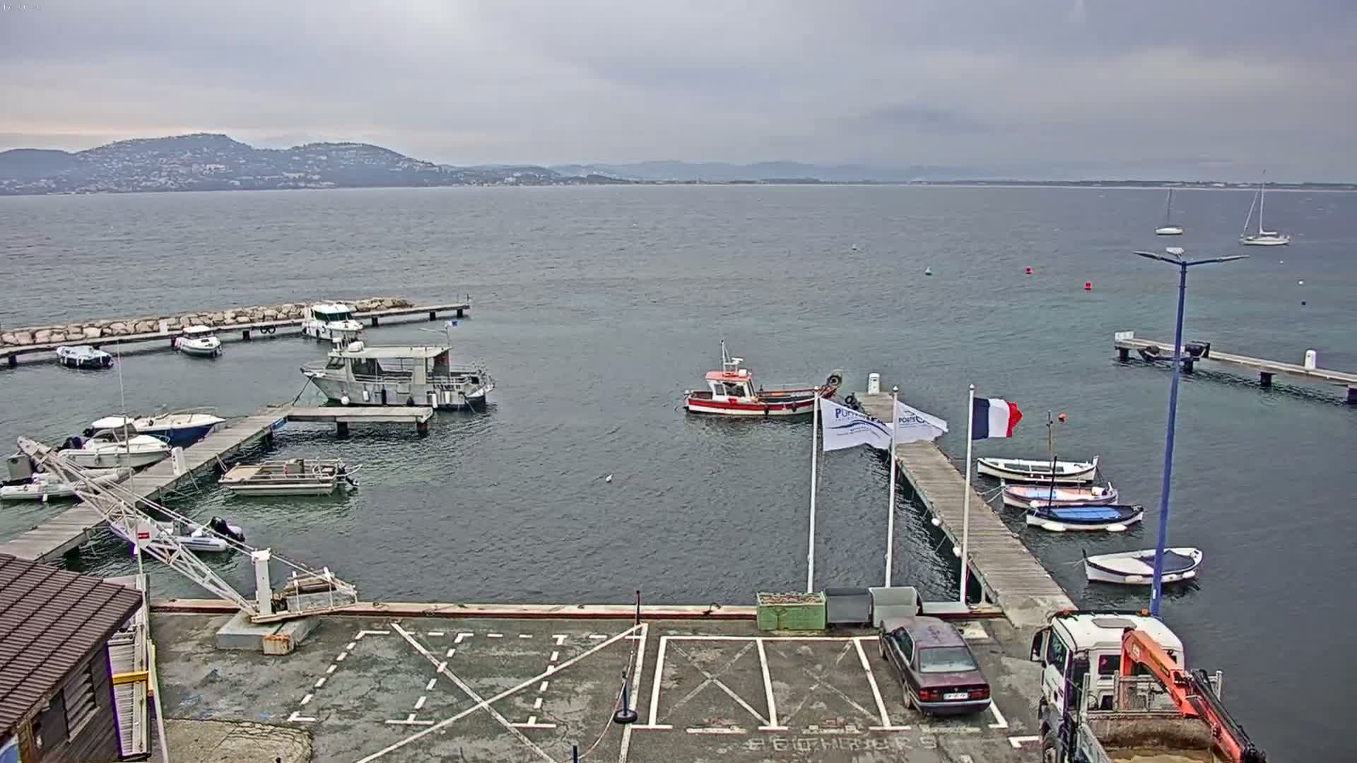 A marina with numerous boats docked at piers, a small parking lot, and a distant mountainous coastline under an overcast sky.