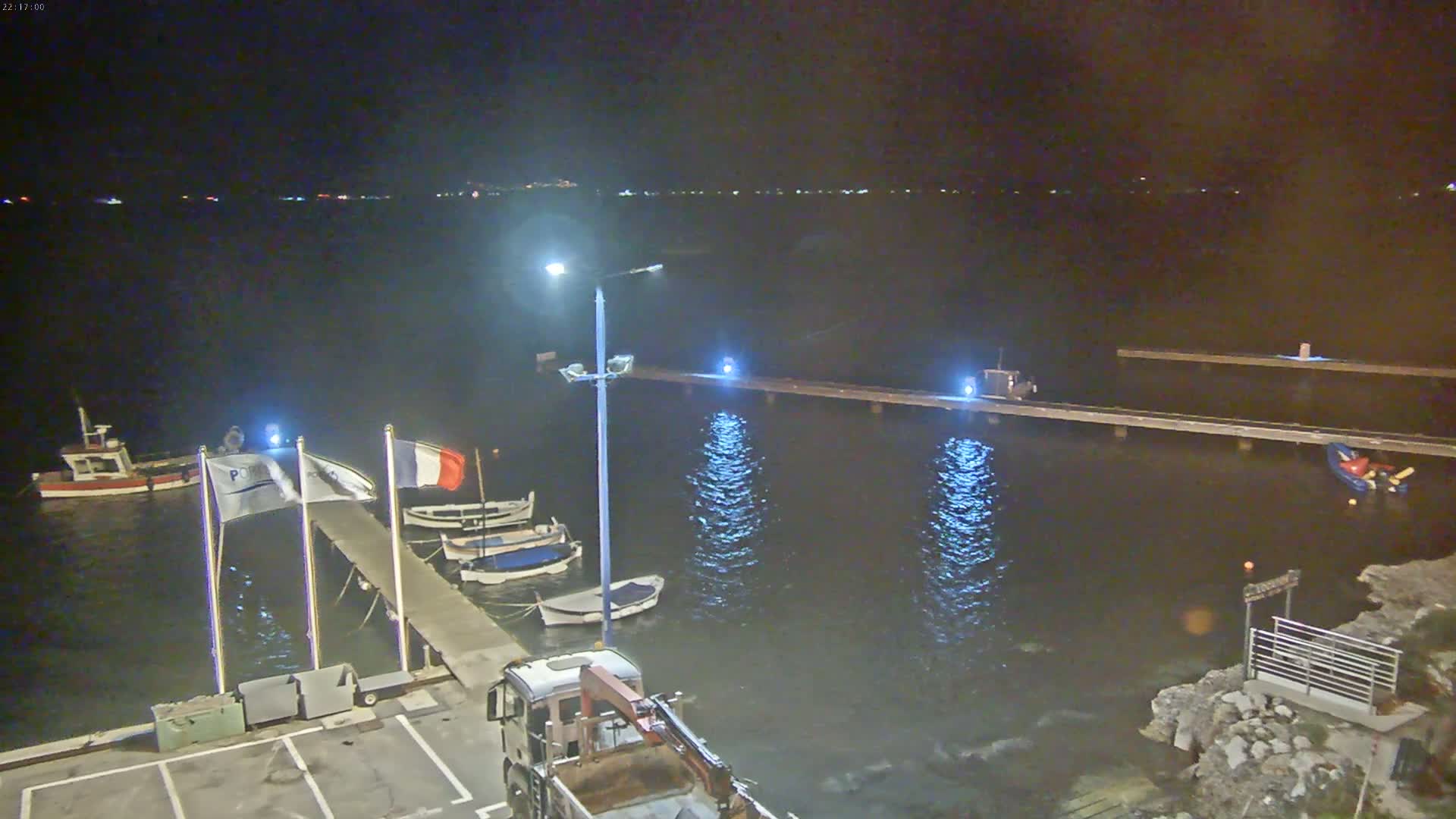 A nighttime view of a calm harbor with several small boats moored at a pier, illuminated by lights, under a dark sky.