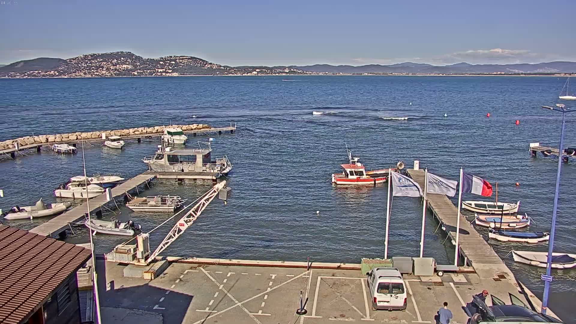 A small marina with numerous boats, a crane, and a parking lot is situated by calm, blue water under a clear, sunny sky with distant hills visible on the horizon.