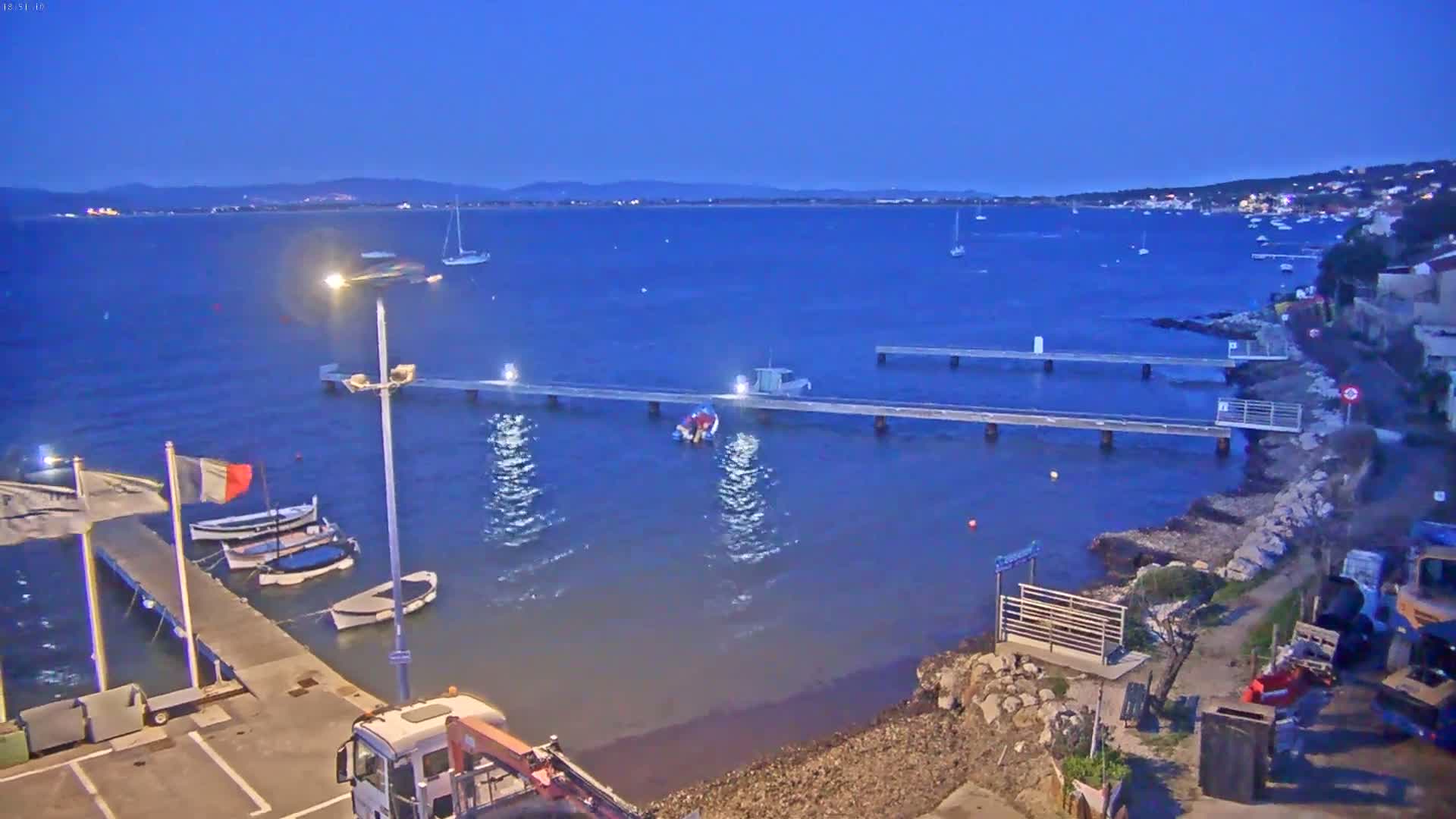 A twilight view of a calm bay with several boats and two piers, one with people on it, under a clear, dark blue sky.