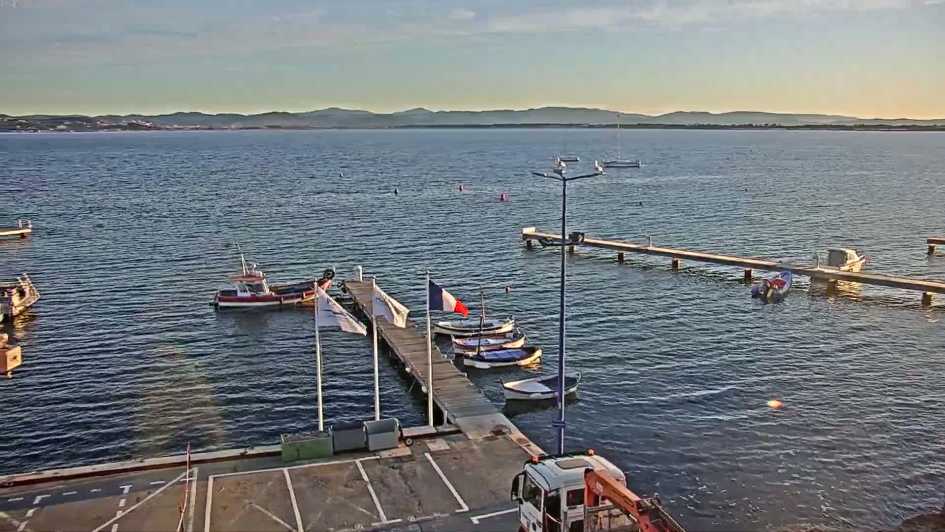 Several small boats are moored at a wooden pier on calm water under a clear, sunny sky, with distant hills visible in the background.