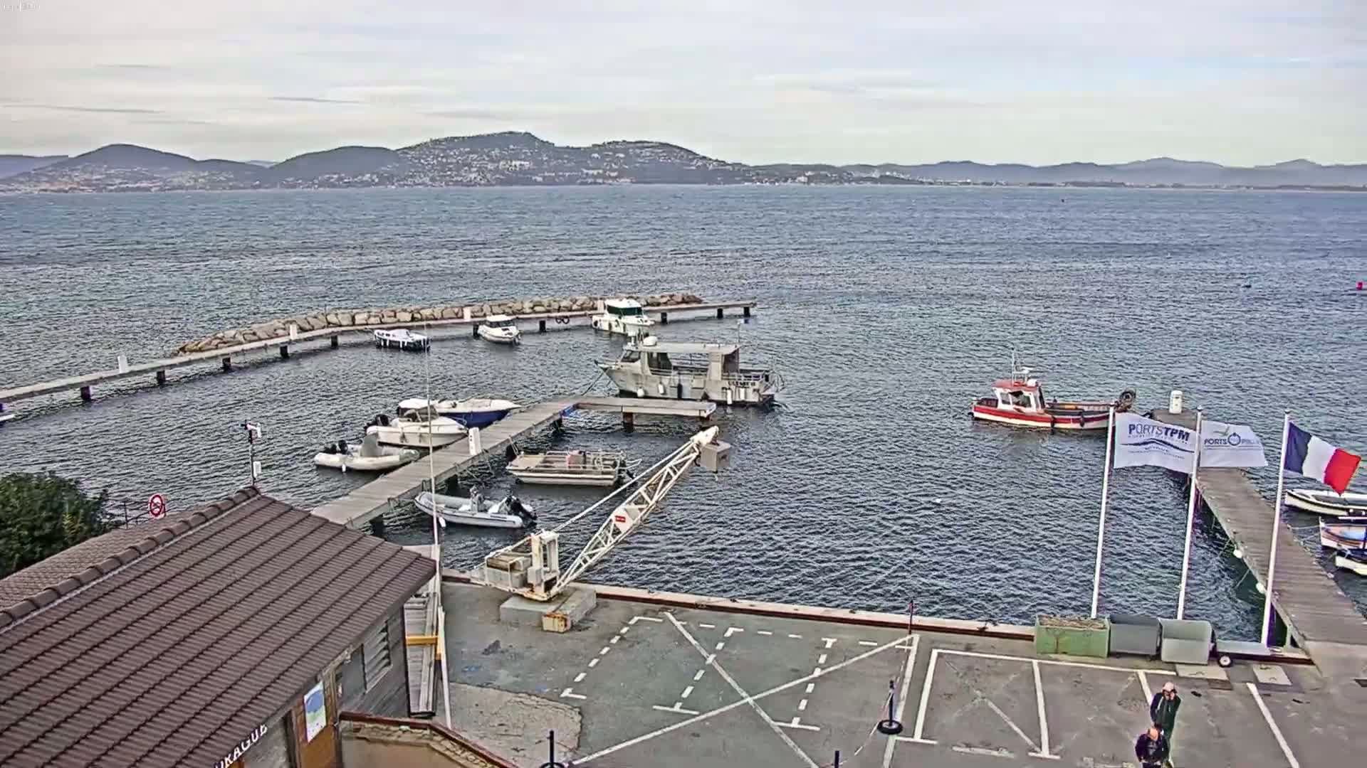 A small marina with numerous boats docked at piers, a crane, and a small building, overlooks a calm body of water with distant mountains under an overcast sky.