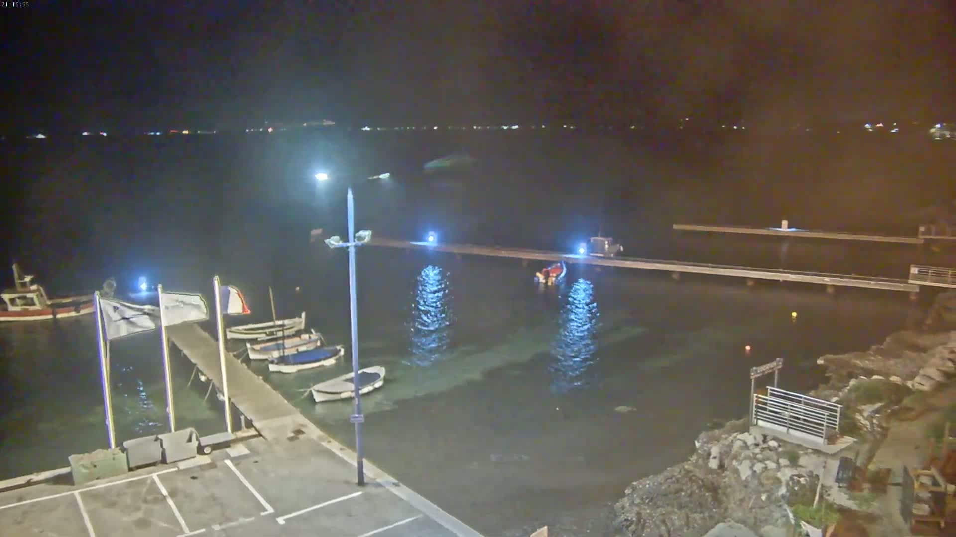 A nighttime view of a small harbor with several boats moored at a pier, illuminated by lights, under a clear, dark sky.