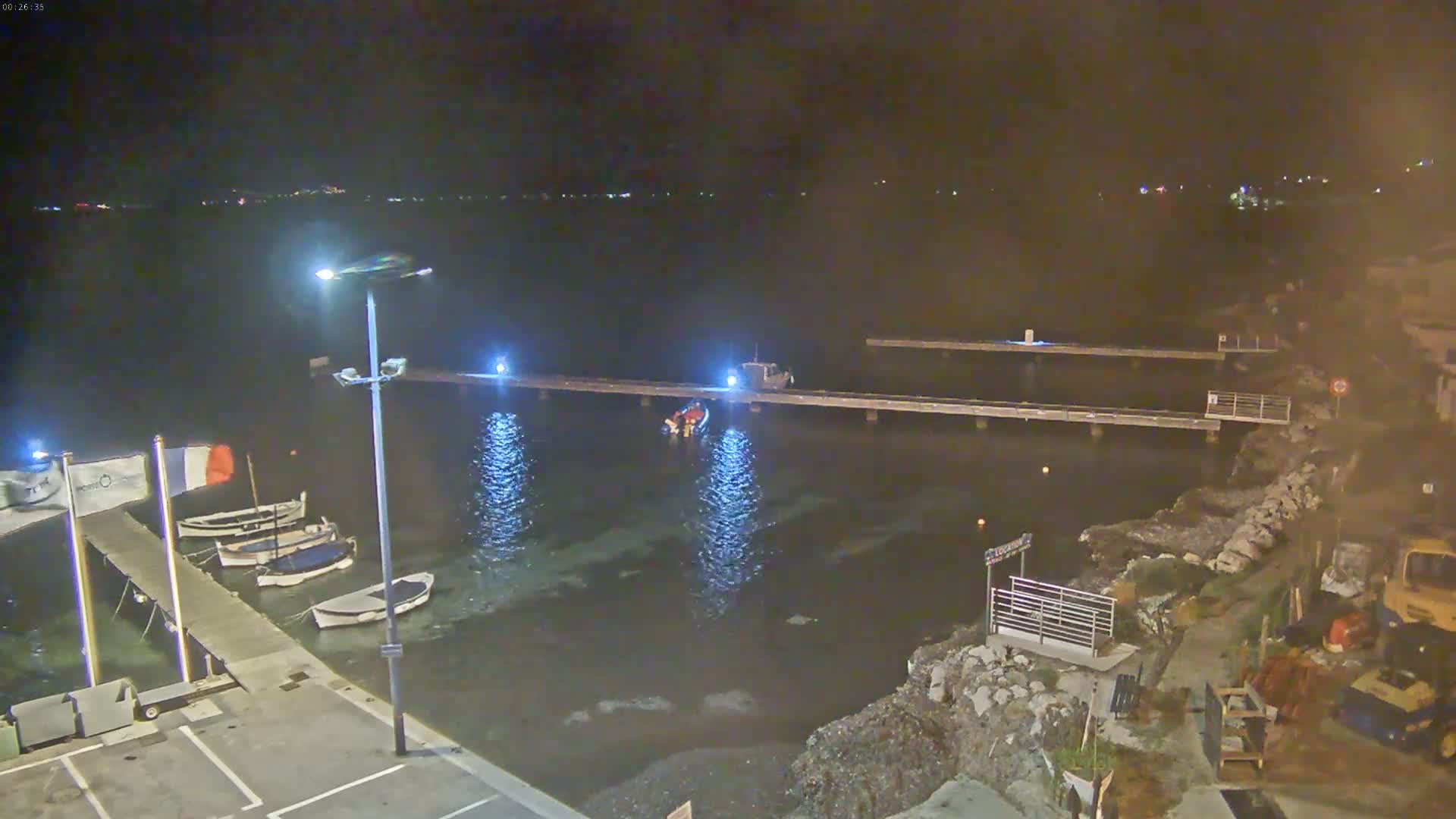 At night, a small harbor with docked boats is illuminated under calm water, with construction equipment visible on the shore.