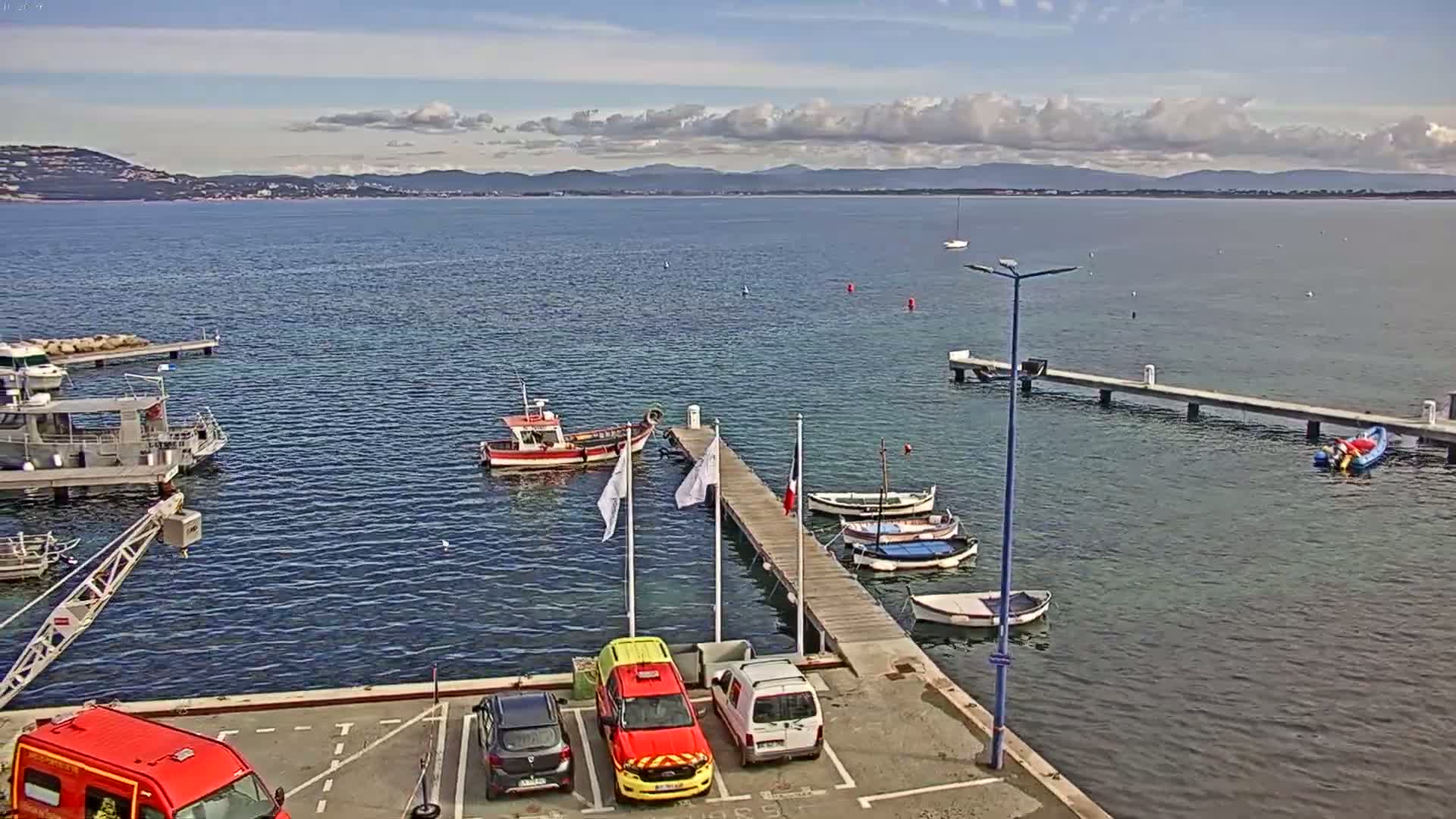 A small harbor with several boats moored at docks, a few cars parked in a lot, and a distant mountainous coastline under partly cloudy skies.