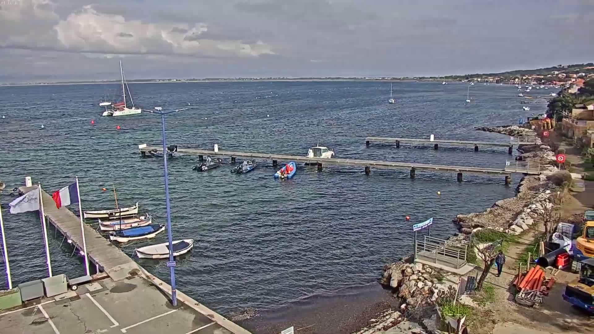 A calm, partly cloudy day shows a harbor with several boats moored at docks, some smaller vessels in the water, and a shoreline with construction equipment.