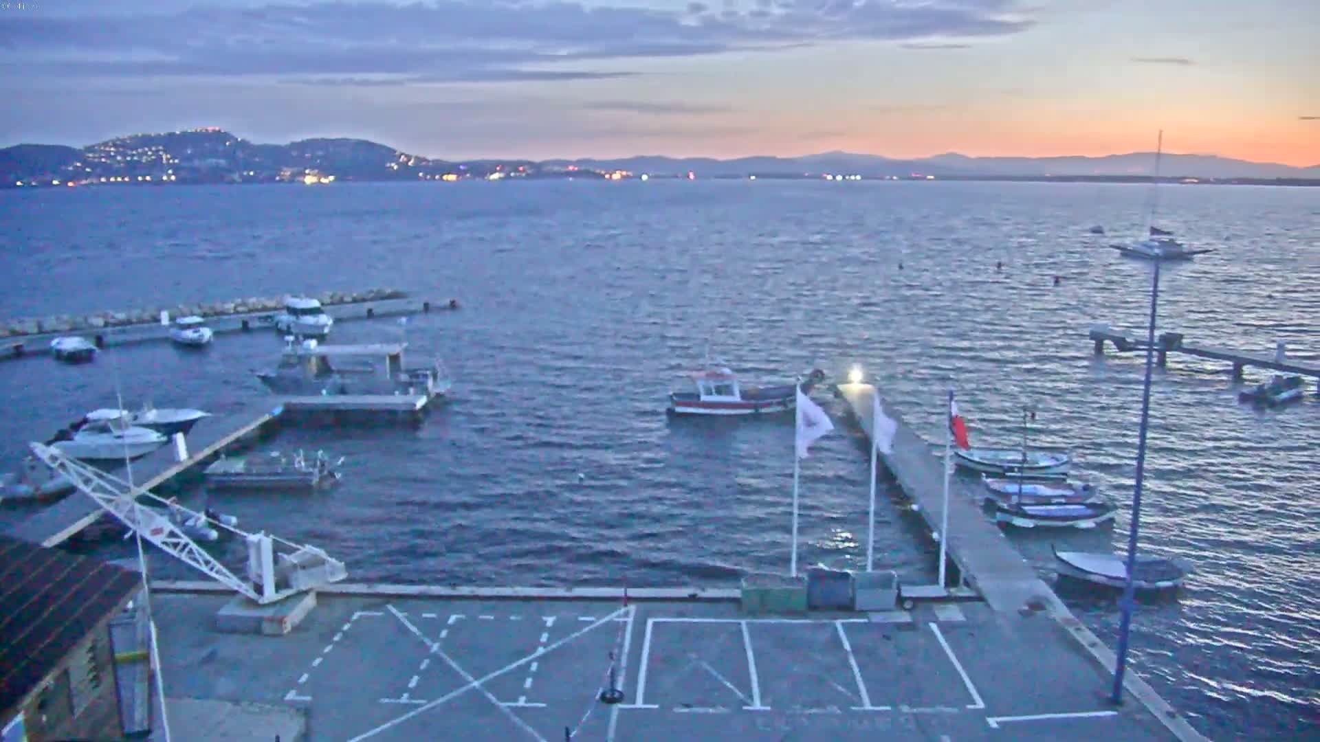 A calm, twilight-lit harbor scene shows numerous boats moored at docks, with a distant, softly lit shoreline visible across the water.