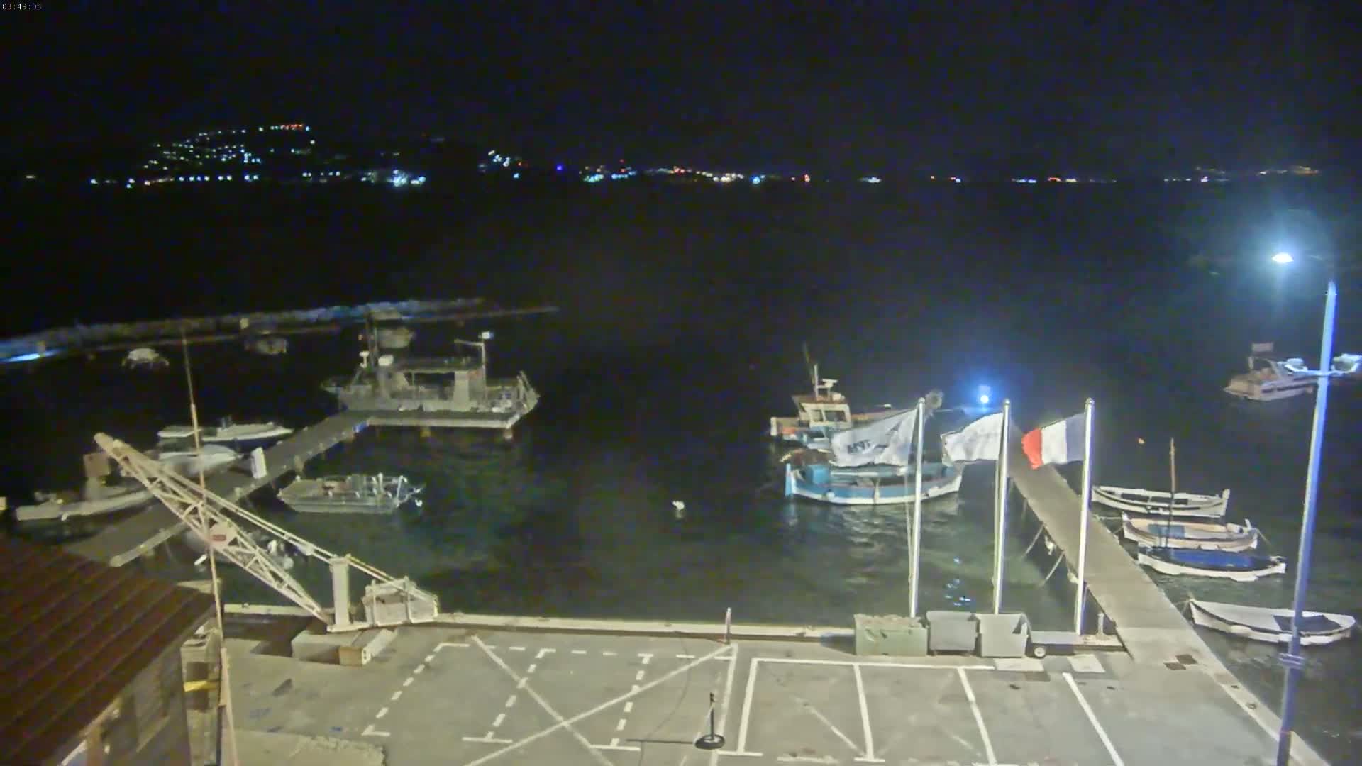 A nighttime view of a harbor with several boats moored at docks, under a clear, dark sky, with a distant town faintly illuminated in the background.