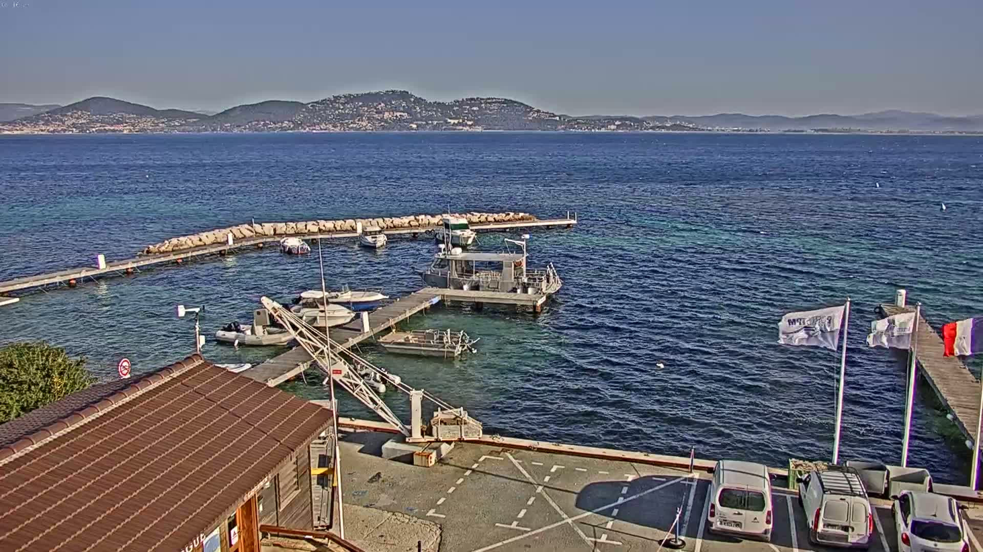 A small marina with several boats docked, a small building, and a few cars parked, overlooks a calm sea with a distant mountain range under a clear, sunny sky.