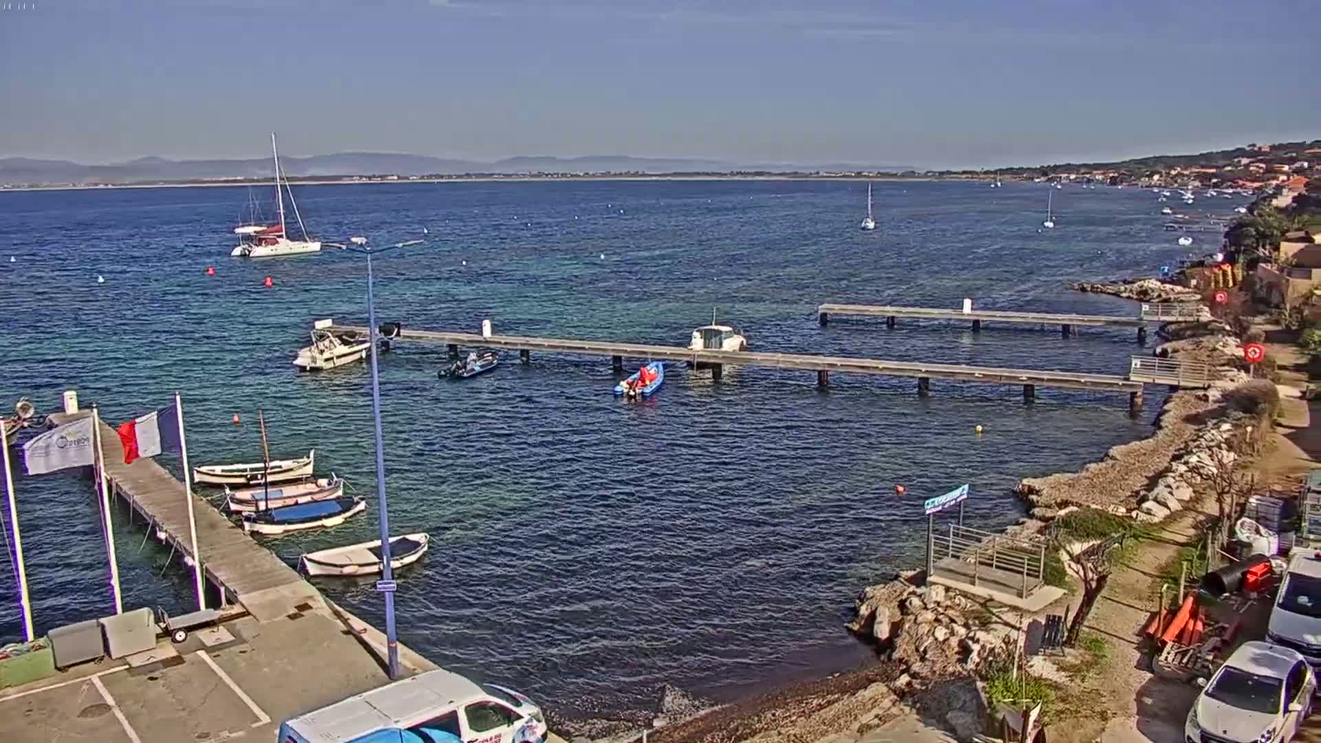 A calm, sunny day reveals a harbor scene with several boats moored at wooden docks extending from a rocky shoreline, and additional boats in the water.