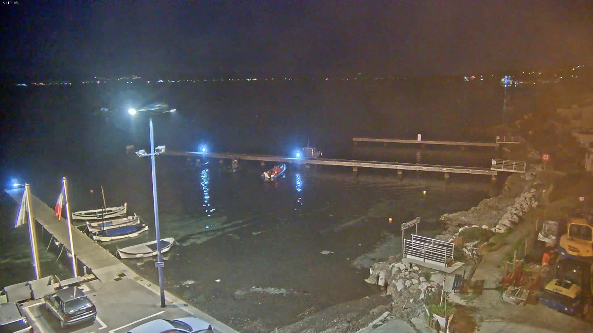 At night, a small motorboat is near a pier with several other boats moored alongside a dark body of water, with construction equipment visible on the shore.