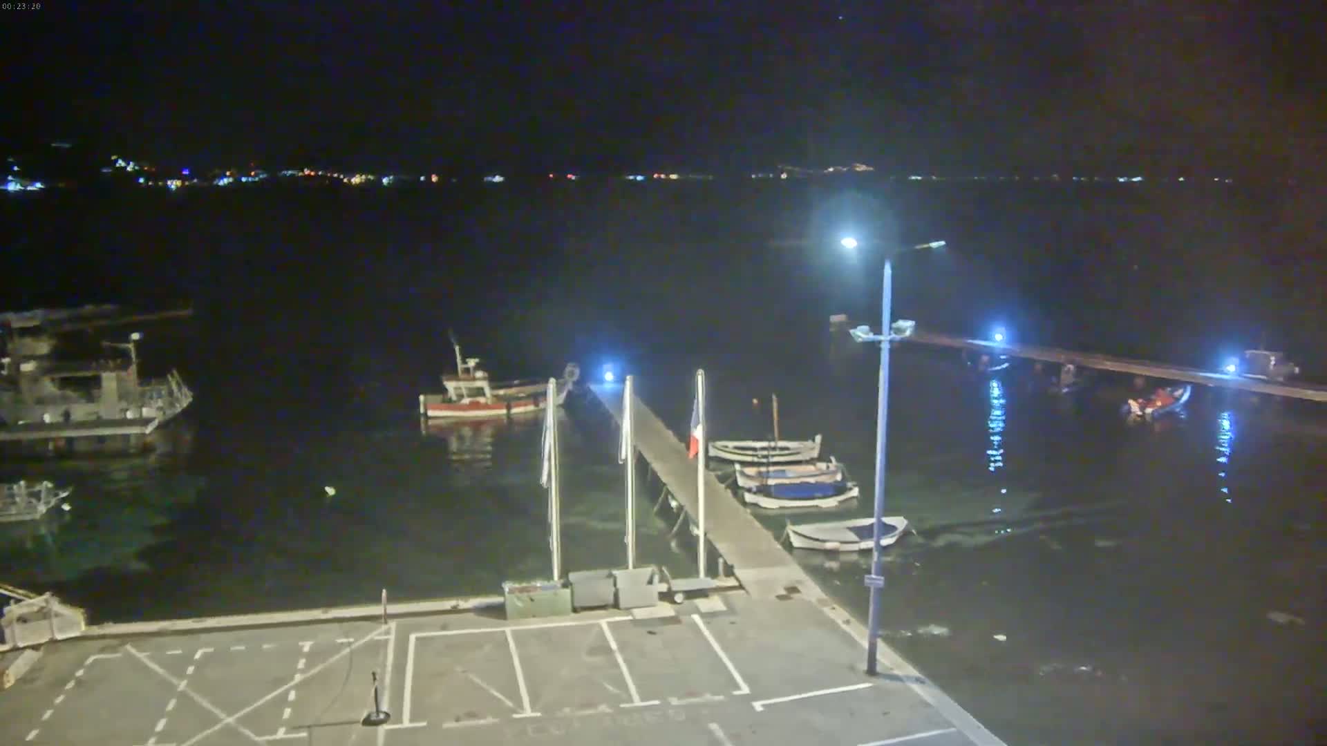 A nighttime, calm harbor scene shows several small boats moored at a pier with a few lights illuminating the water and a distant town's lights visible on the horizon.