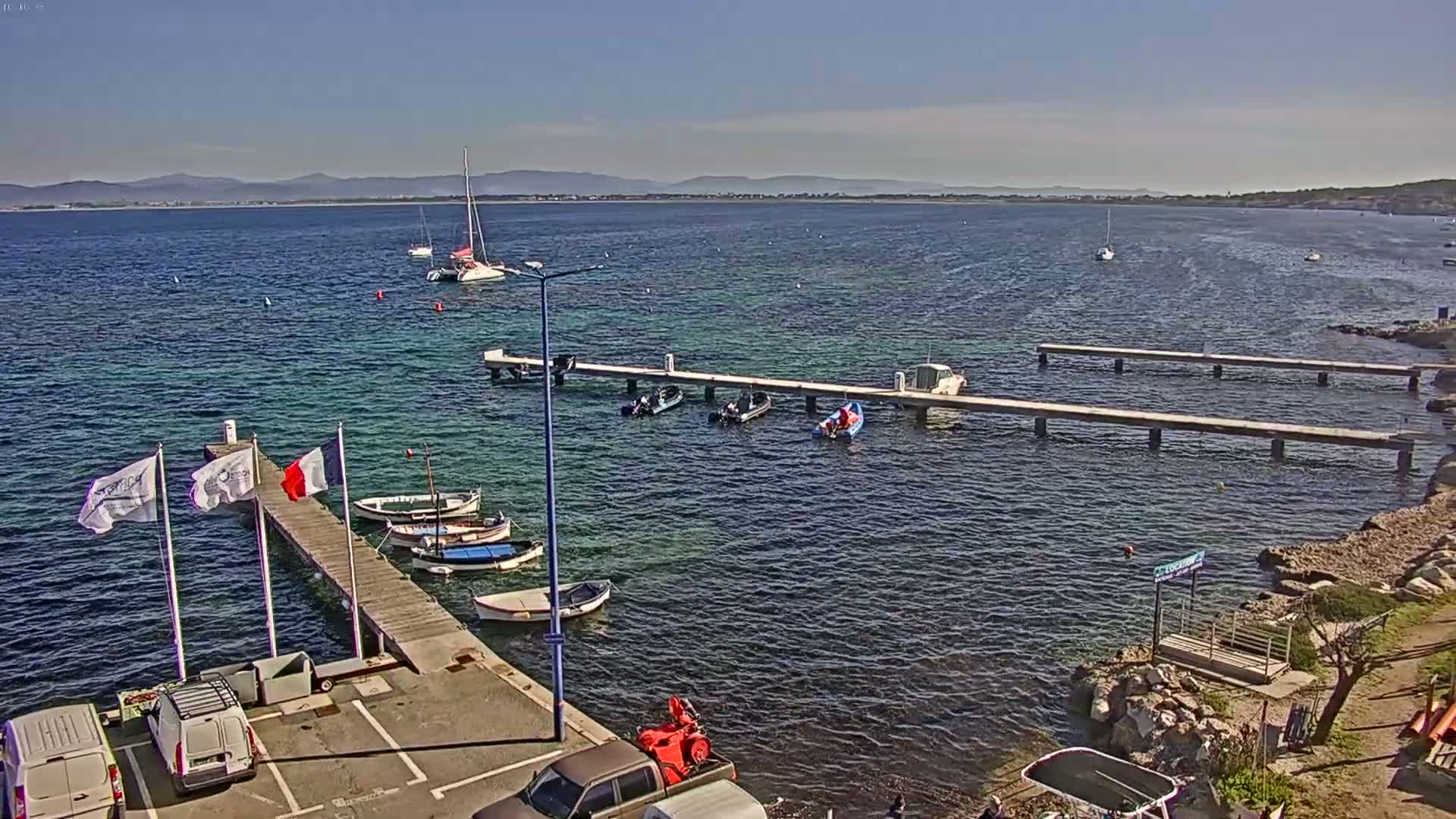 A calm, sunny day shows a harbor with several boats docked at wooden piers, with additional boats and sailboats in the clear water and distant mountains visible on the horizon.
