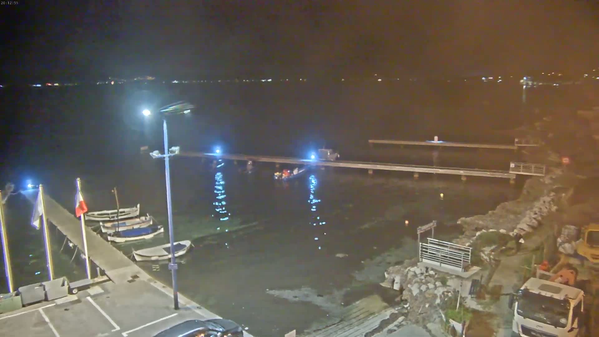 A nighttime view of a harbor with several small boats moored at a pier, under calm conditions, with construction vehicles and workers visible on land.