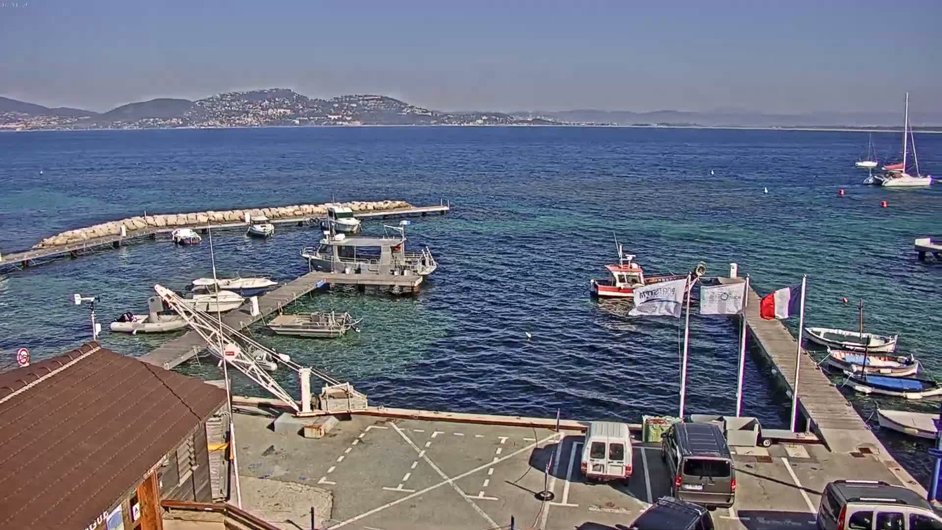 A small marina with several boats and a few cars in a parking lot is nestled against a calm, blue sea with distant mountains under a clear, sunny sky.