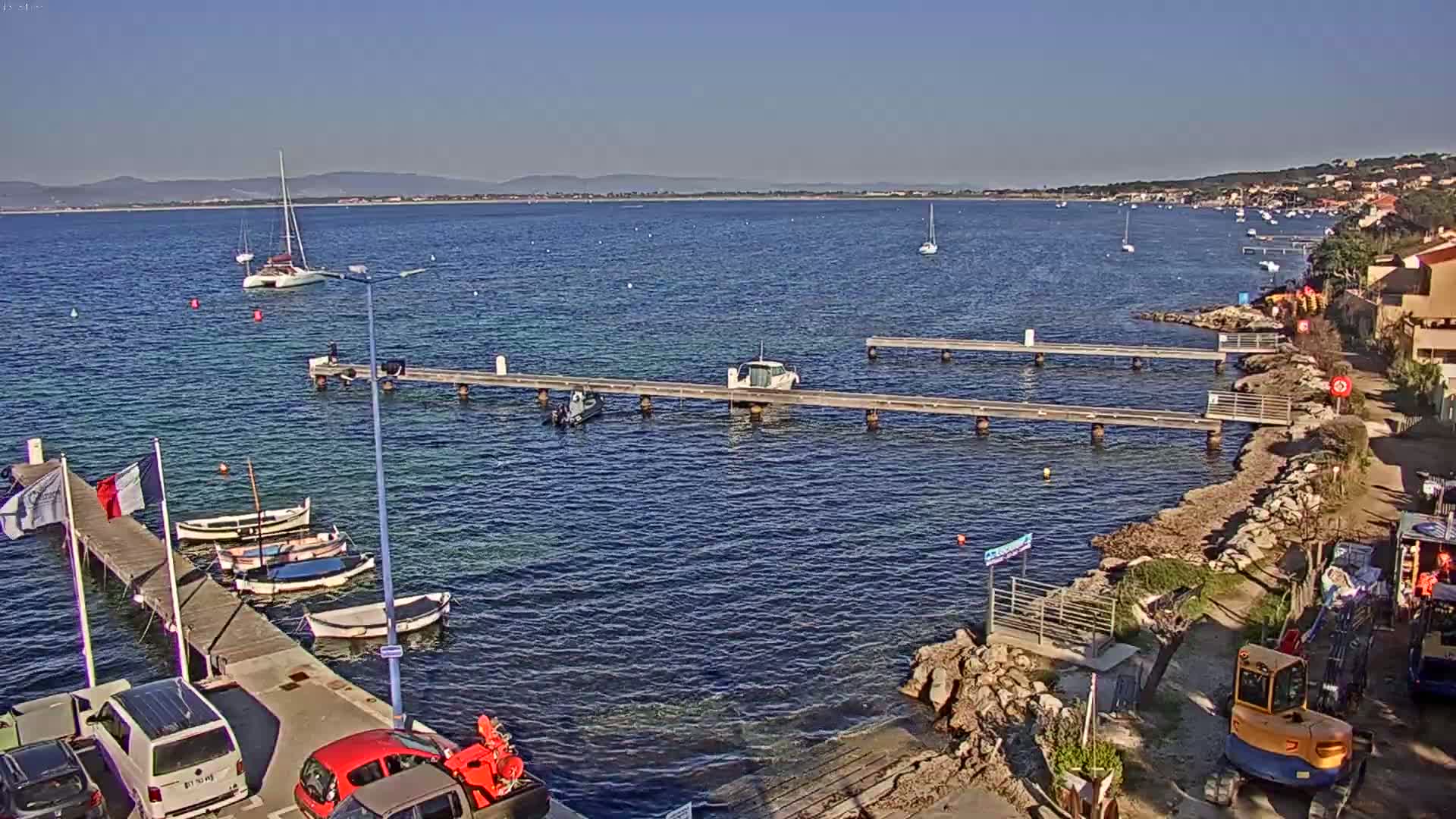 A clear, sunny day shows a calm body of water with several boats moored at wooden piers along a rocky shoreline, with additional boats and vehicles visible along the coast.