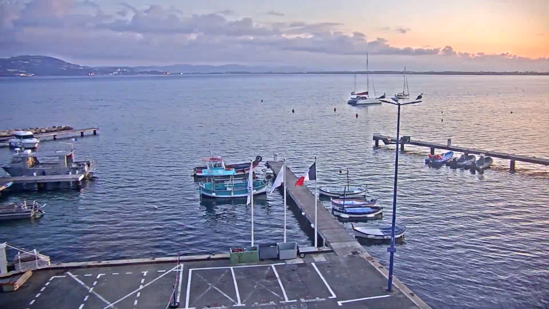 A calm harbor at sunset with several boats docked at piers, and additional boats and sailboats anchored in the water.