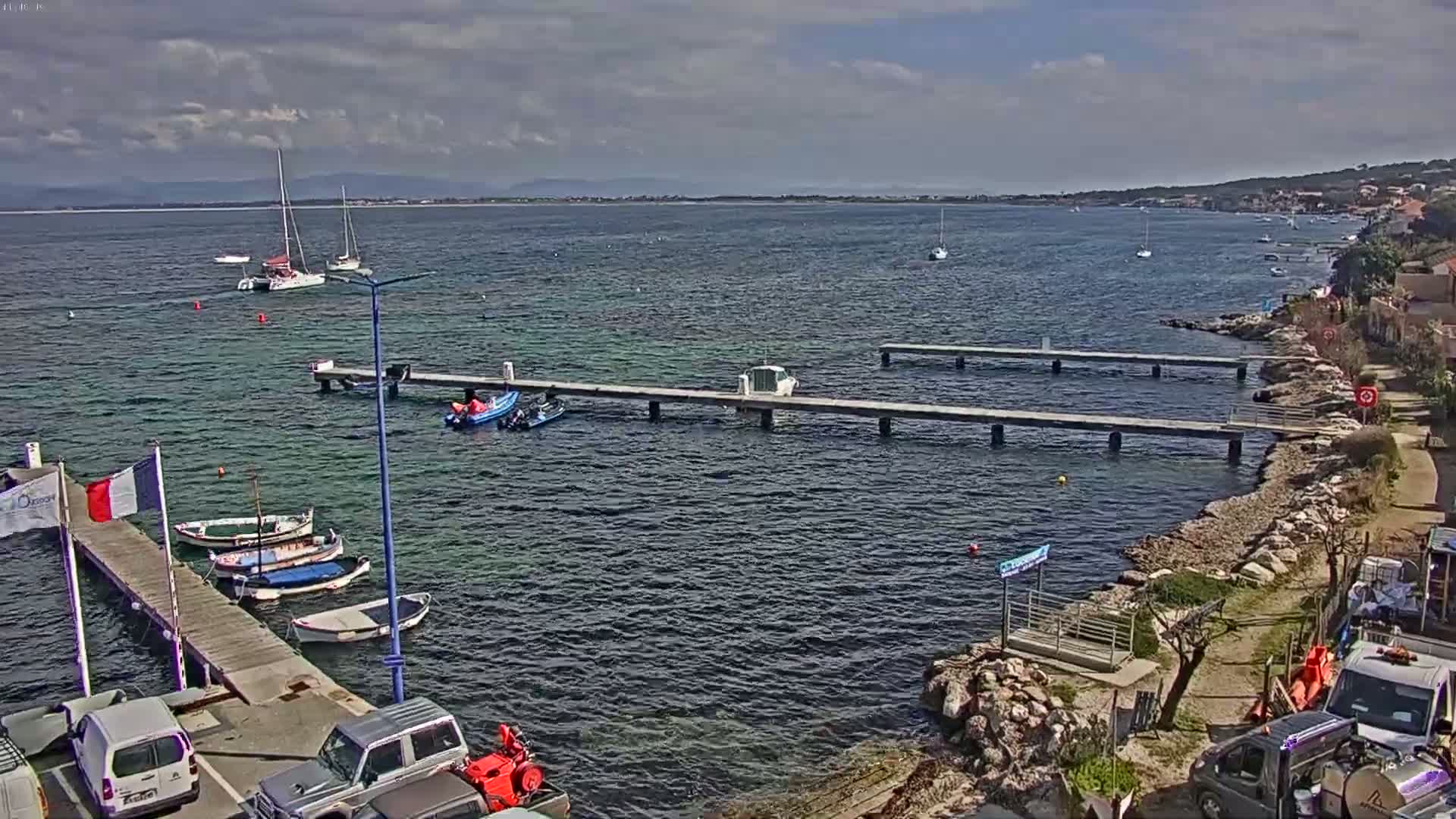 A partly cloudy day shows a calm body of water with several boats moored at a dock, other boats sailing in the distance, and a rocky shoreline with vehicles parked nearby.
