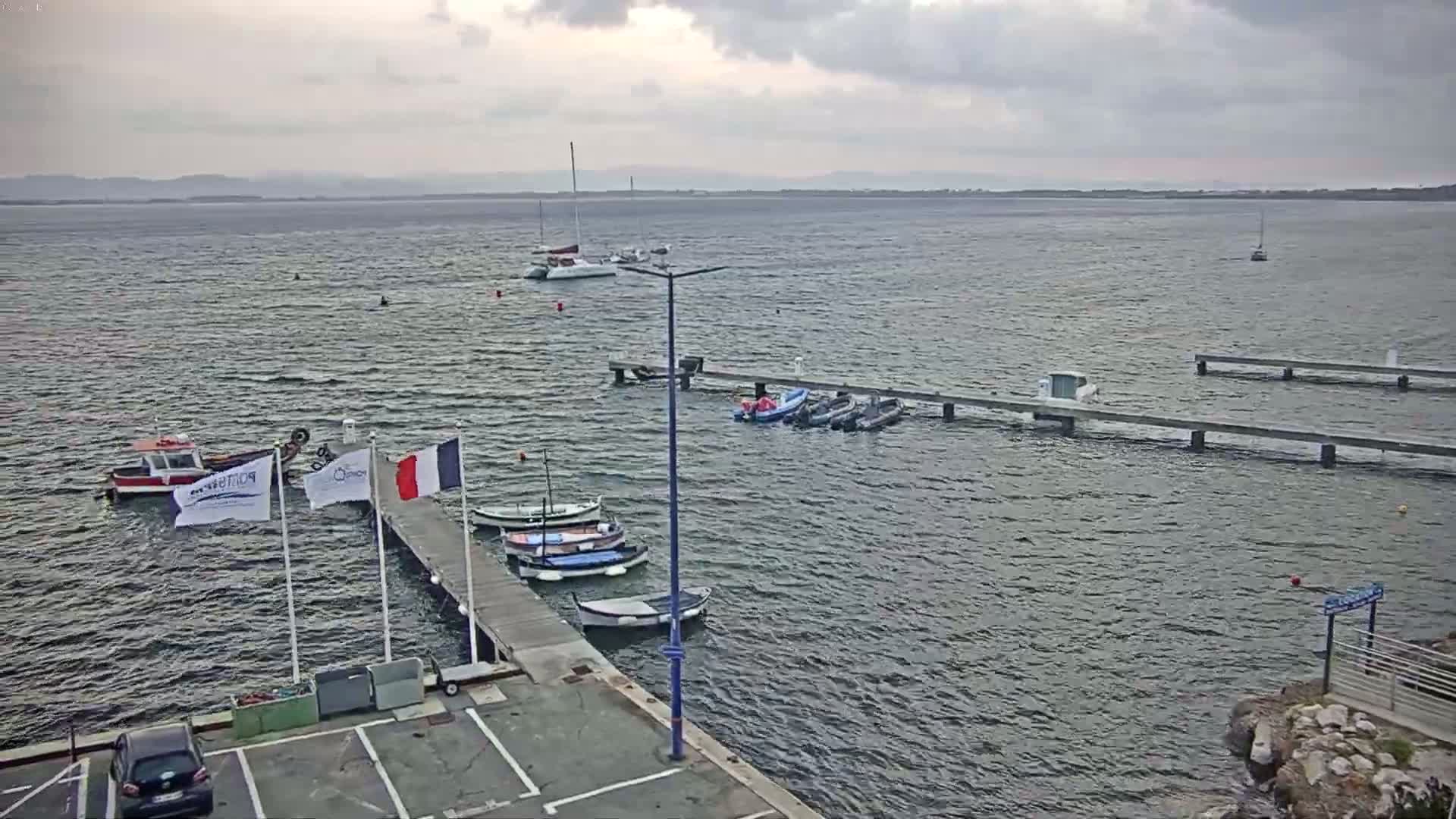 Several boats are moored at a pier on a somewhat choppy body of water under a cloudy sky.
