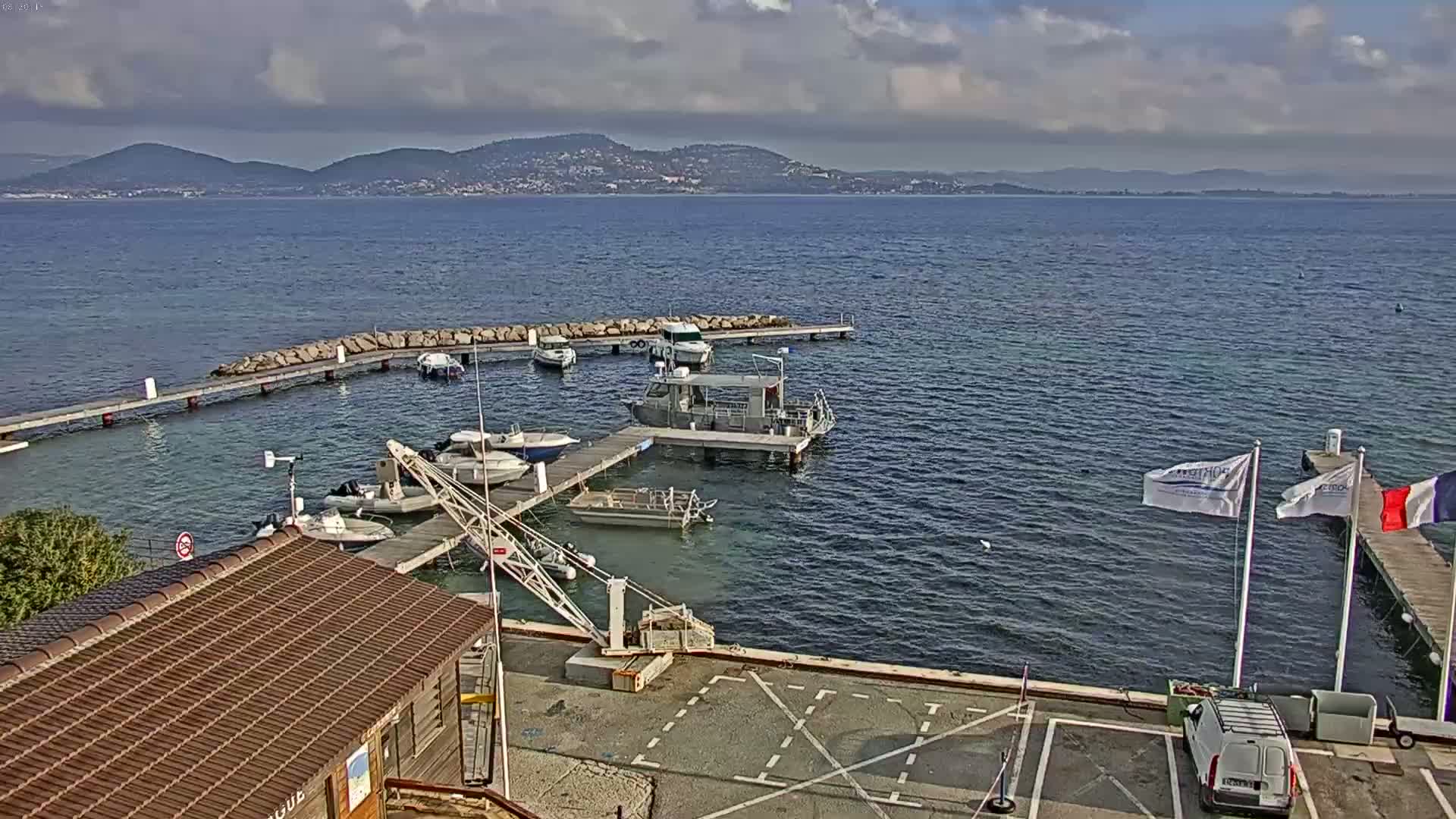 A small marina with several boats docked at piers, a crane, and a small building, overlooks a calm body of water with mountains in the distance under a partly cloudy sky.