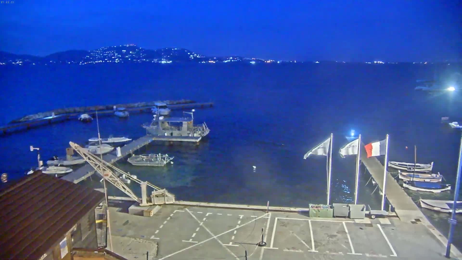 A small marina at twilight, with numerous boats moored at docks, overlooks a calm, dark blue sea under a clear night sky, and a distant, faintly lit shoreline is visible.
